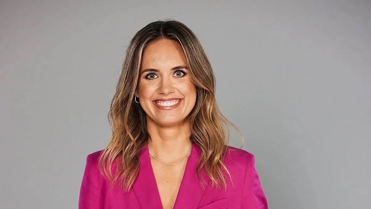 Portrait of a smiling woman with shoulder-length wavy hair, wearing a pink blazer and a delicate necklace, standing against a plain gray background.