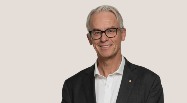 Portrait of a smiling older man with gray hair, glasses, wearing a dark suit and white shirt, standing against a plain background.