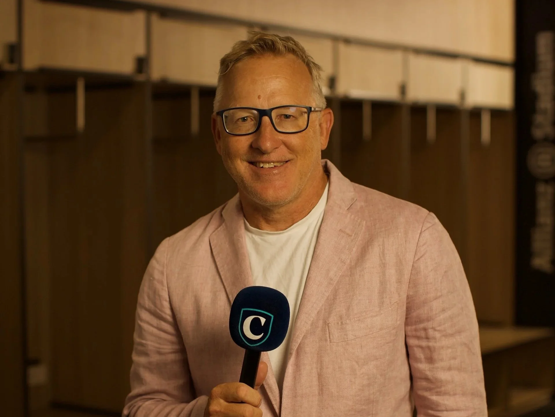 A smiling middle-aged man with gray hair and glasses, wearing a light pink blazer and a white shirt, holding a microphone with a blue 'C' logo, standing indoors with wooden shelves in the background.