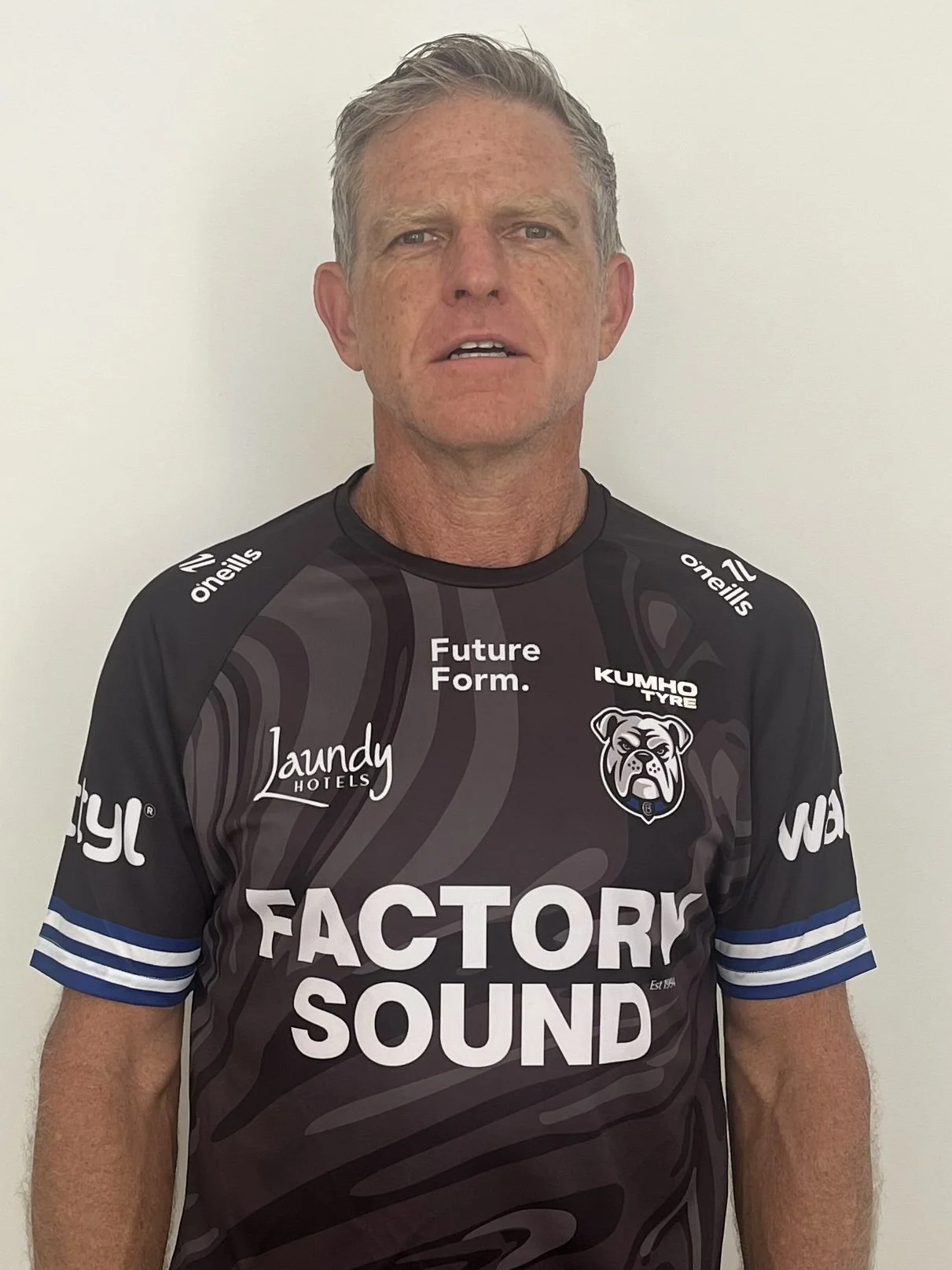 A man in a black and gray sports jersey with blue and white striped sleeves, standing against a plain white wall.