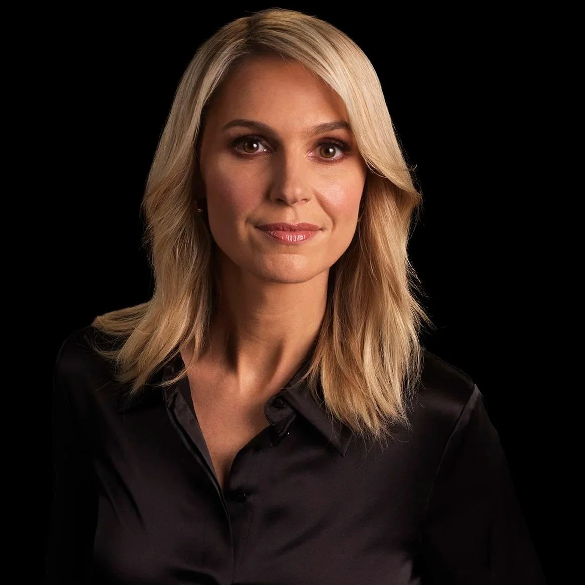 A woman with shoulder-length blonde hair, wearing a black shirt, looking directly at the camera with a slight smile, against a black background.