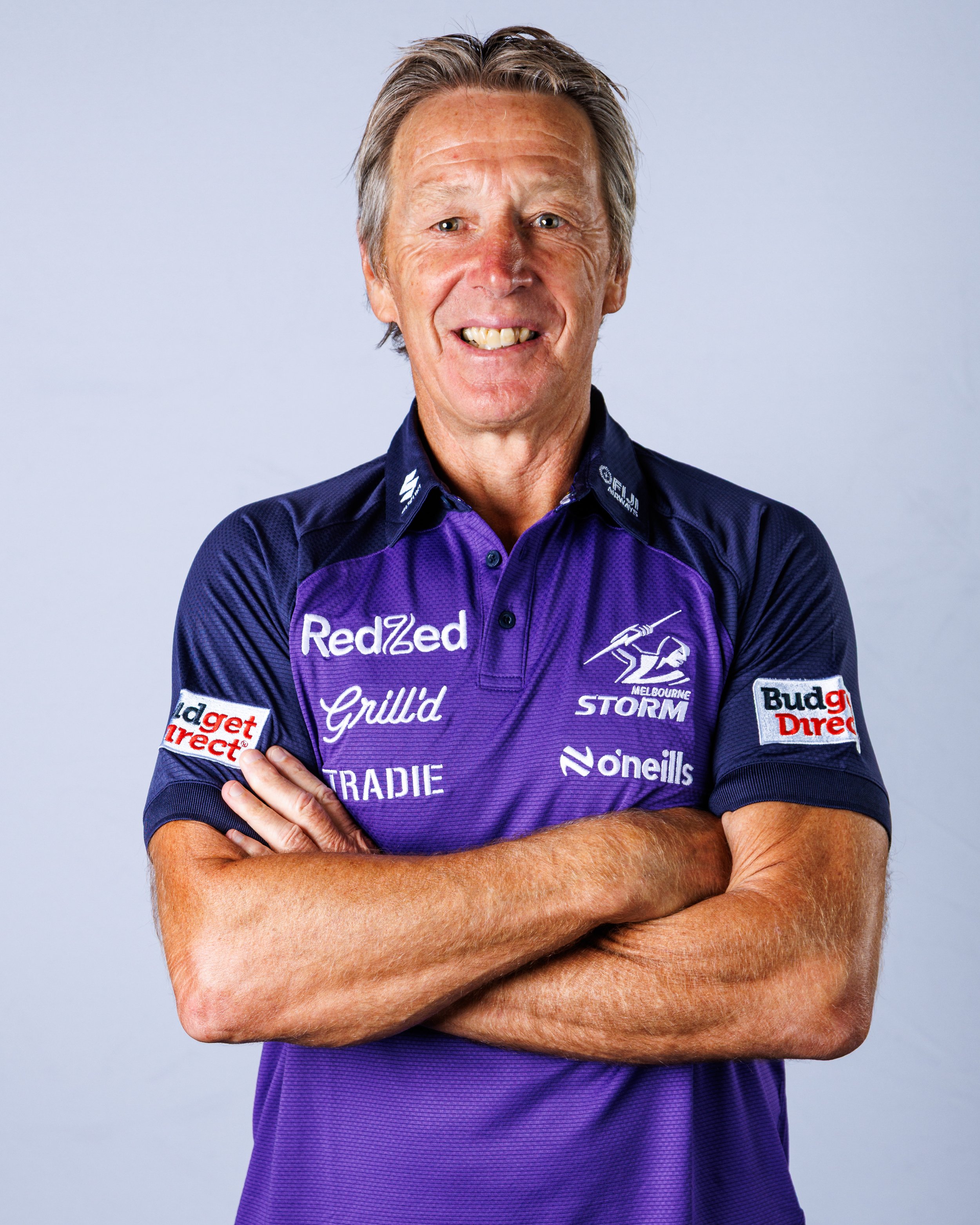 A middle-aged man with gray hair smiling, standing with arms crossed, wearing a purple sports jersey with team and sponsor logos, against a light gray background.