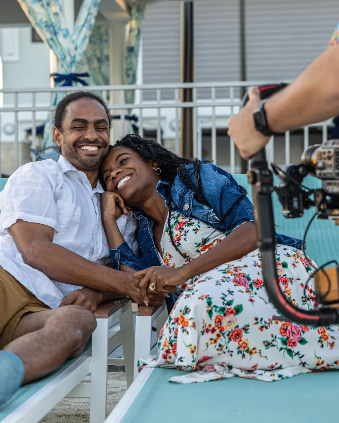 A couple sitting on a bench, smiling and holding hands, while being filmed or photographed by a person using a professional camera.