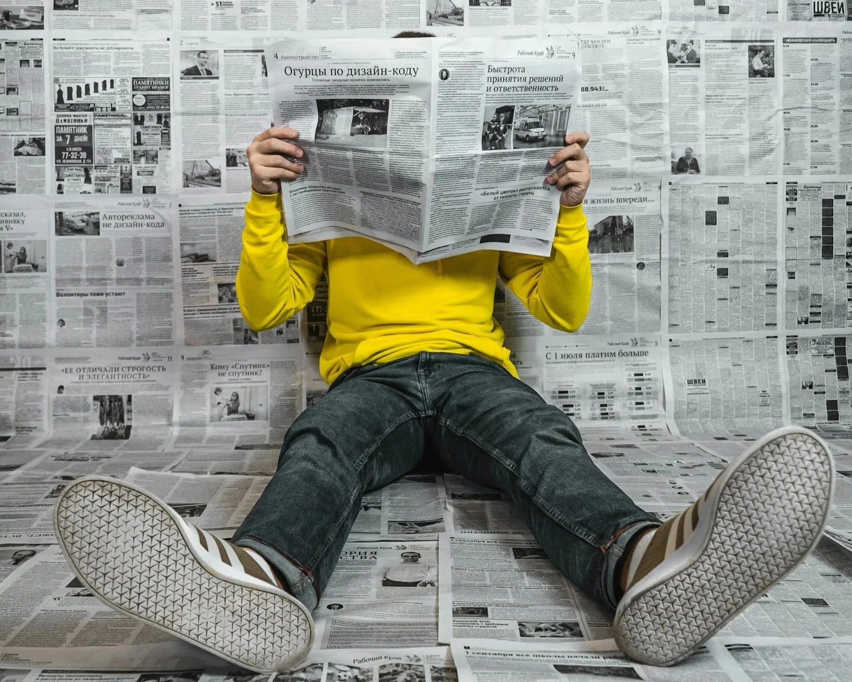Person sitting on the floor against a wall covered with newspapers, wearing a yellow shirt, black jeans, and sneakers, holding and reading a newspaper.