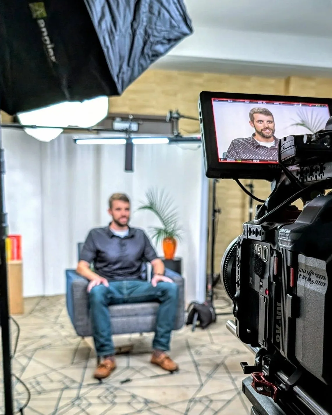 A man sitting on a gray armchair during an interview or recording session, with a camera capturing him and a backdrop of light curtains, a potted plant, and professional lighting equipment in a studio setting.