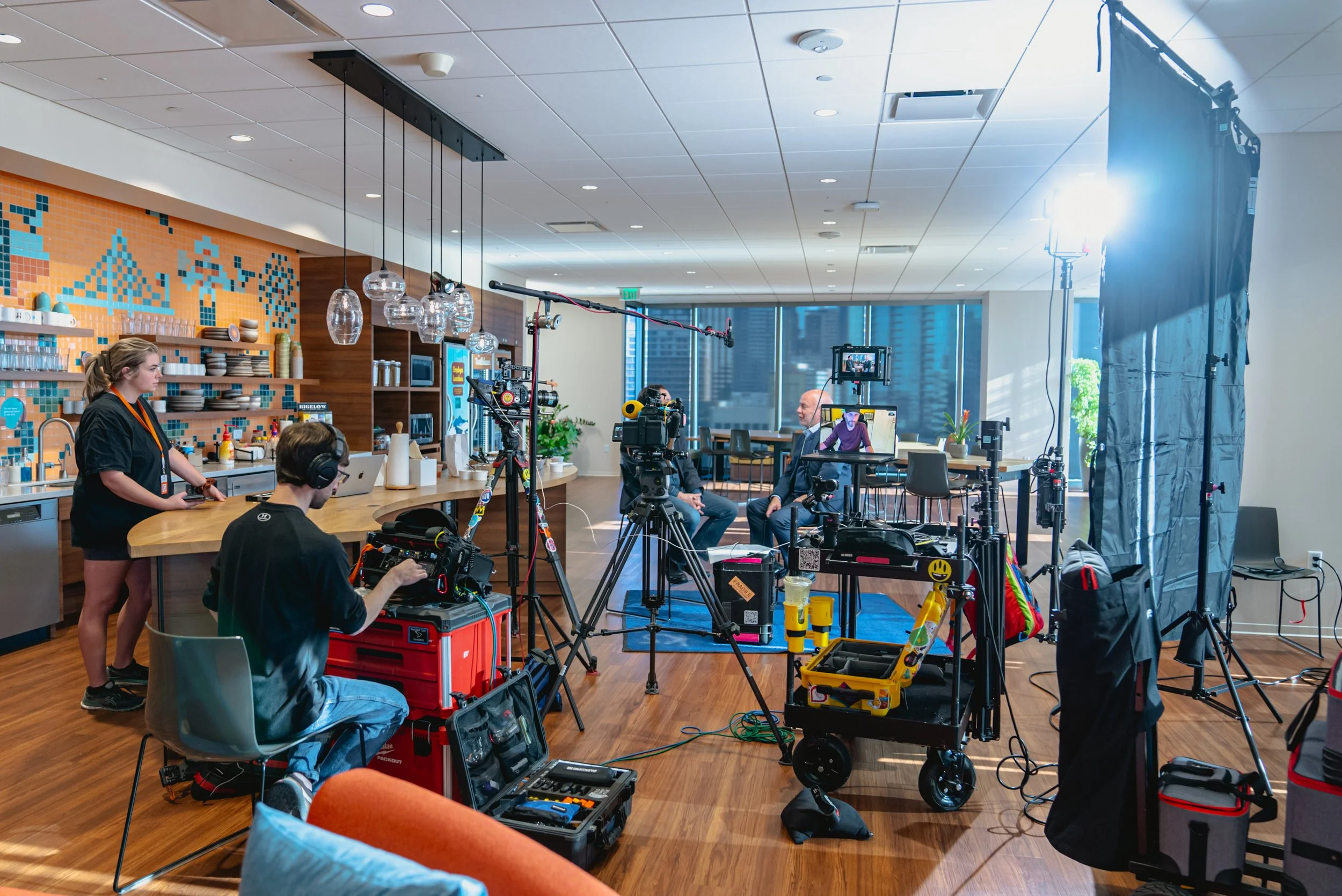 Studio setup with cameras, lighting, and crew members filming an interview with two men in suits sitting across from each other in a room with large windows.