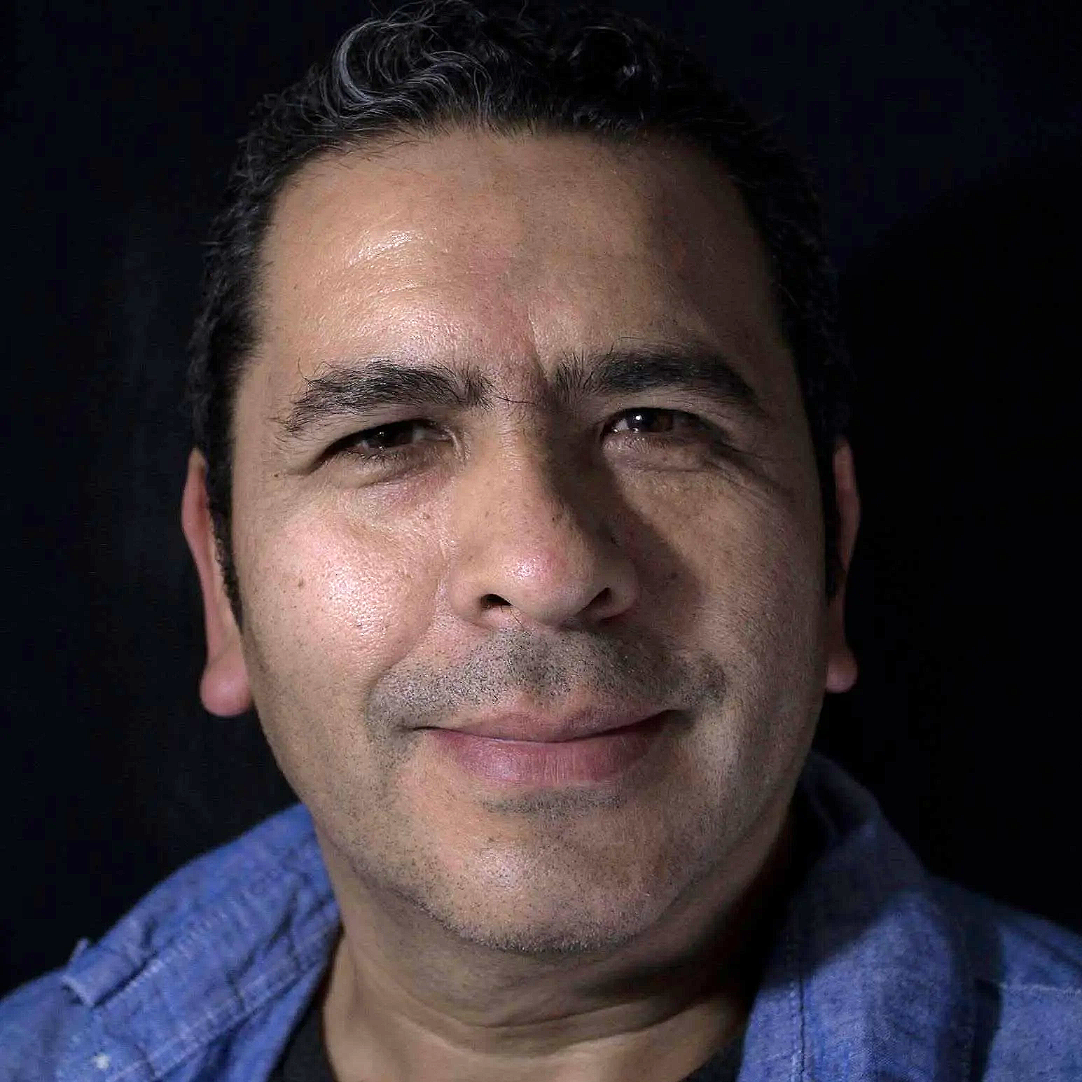 Close-up of a middle-aged man with black hair, slightly wavy, smiling softly, wearing a blue shirt against a dark background.