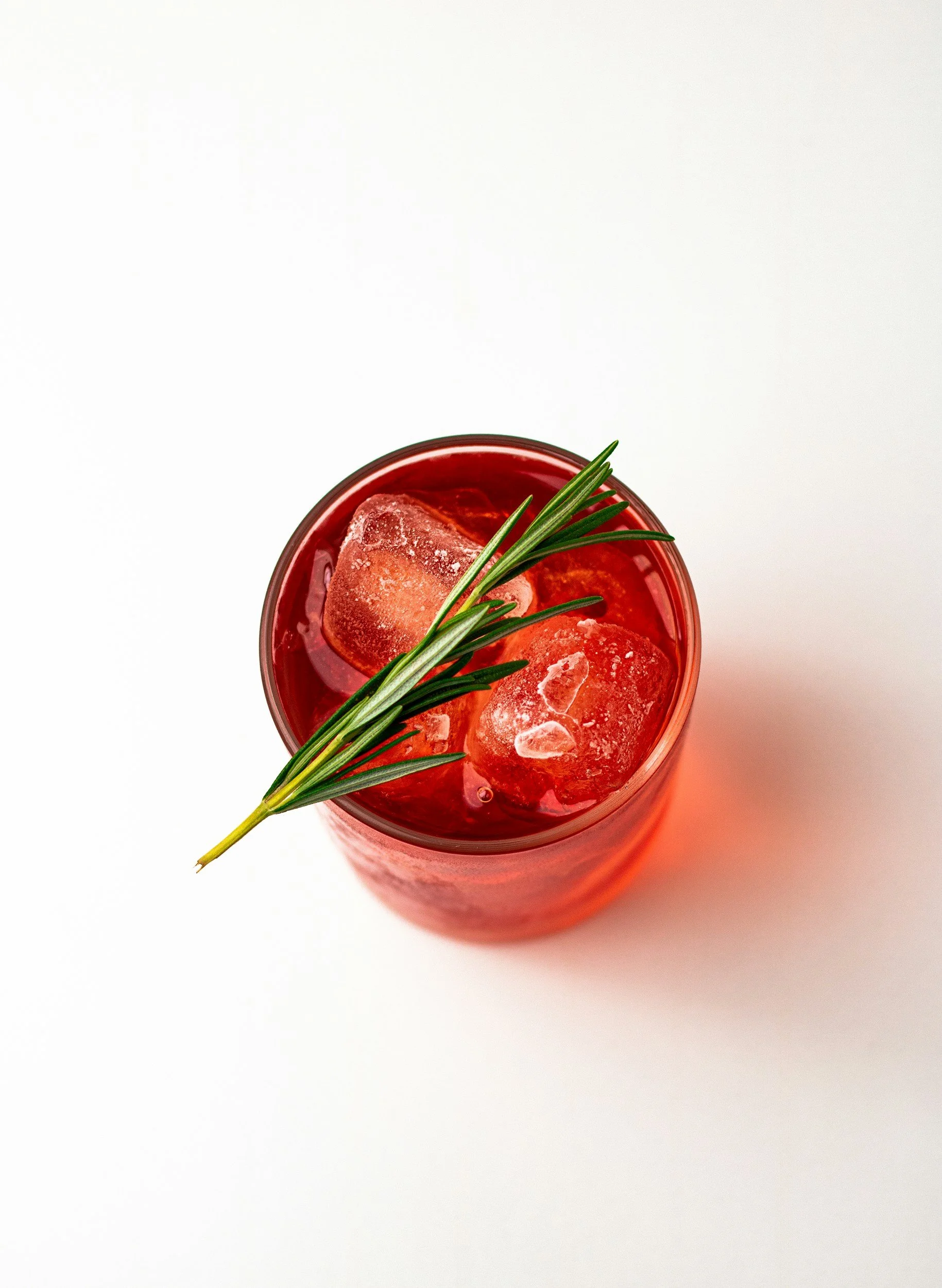 Red beverage in a glass with ice cubes and a sprig of rosemary, on a white surface