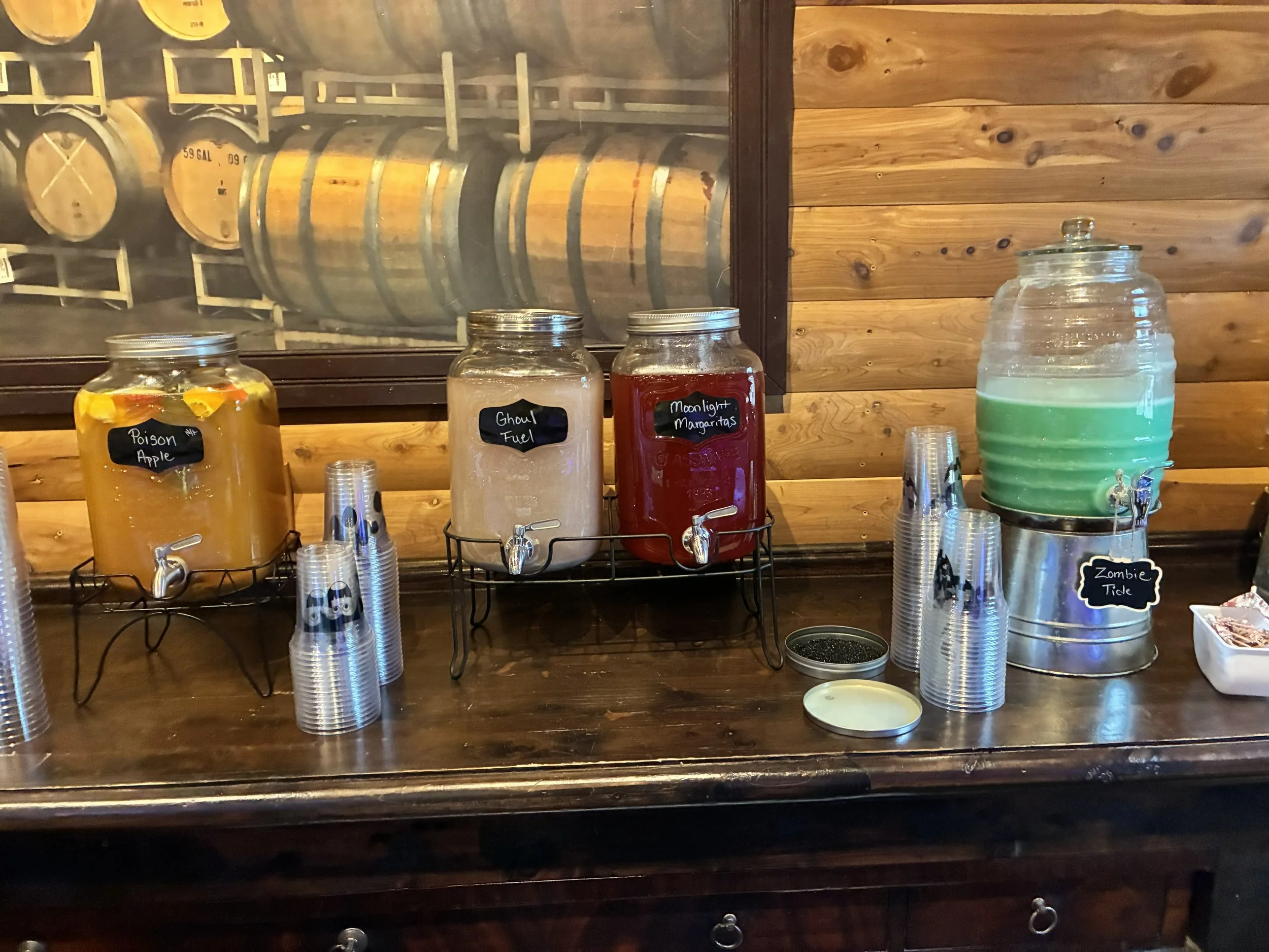 Drink dispensers labeled 'Poison Apple', 'Ghoul Fuel', 'Moonlight Margaritas', and 'Zombie Tide' on a wooden bar counter with stacks of plastic cups and paper napkins, against a wooden wall with a window showing barrels inside a storage room.