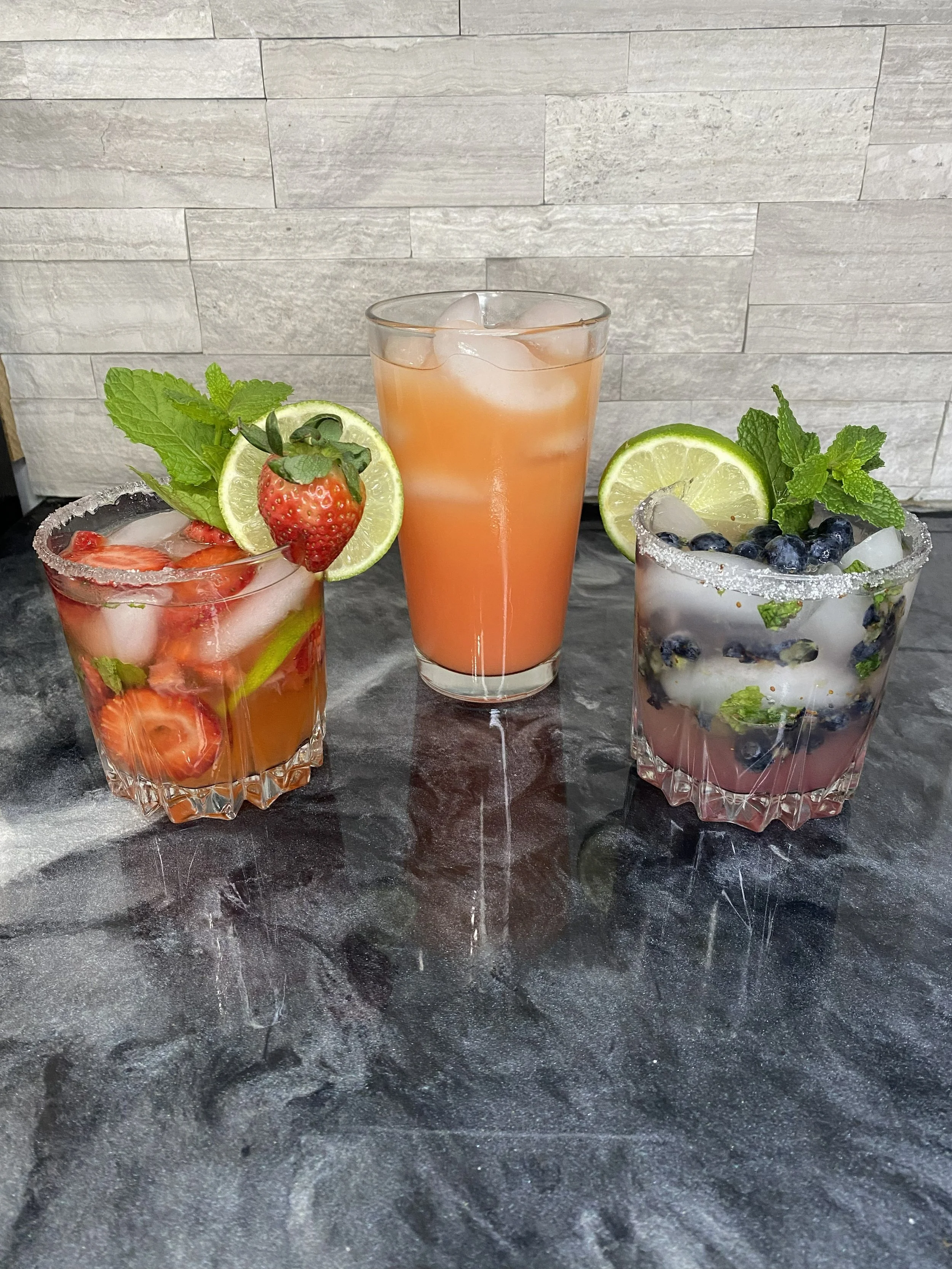 Three cocktails on a dark marble table, garnished with fresh fruit and mint, against a light gray stone wall background.