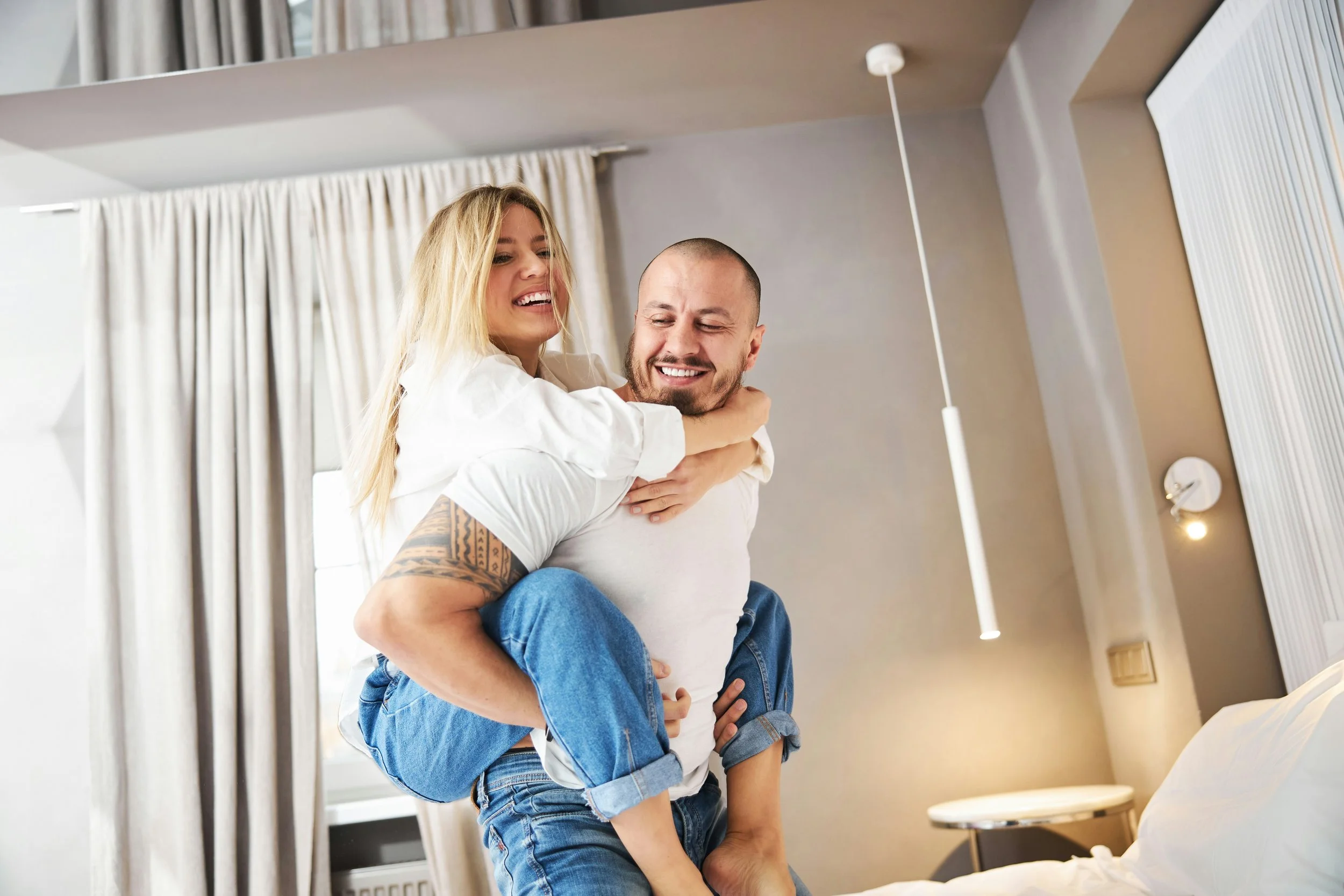 A happy couple in a bedroom, with the woman on the man's back, smiling and laughing.