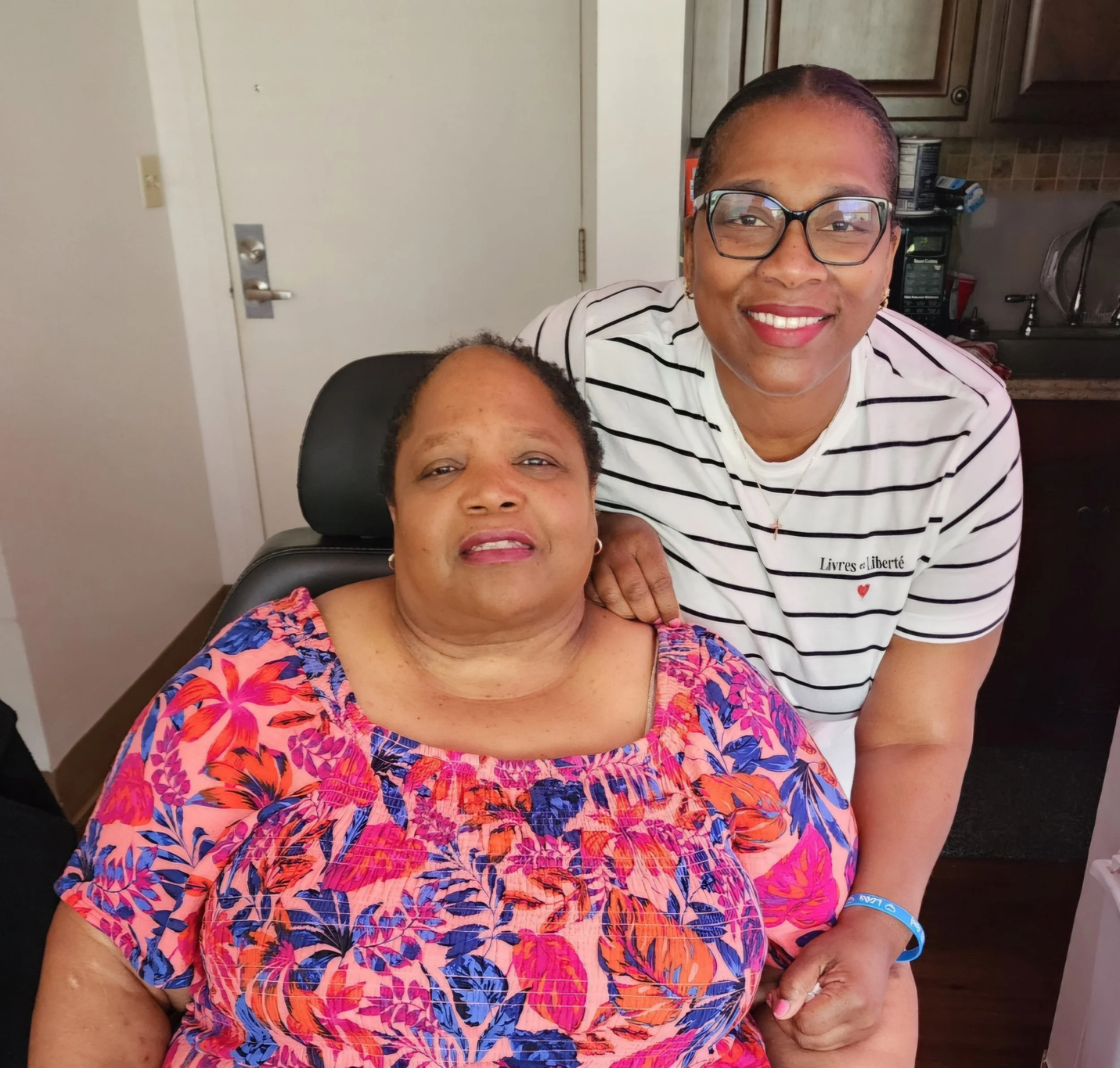 Two women, one sitting in a wheelchair and the other standing behind her, smiling in a kitchen.
