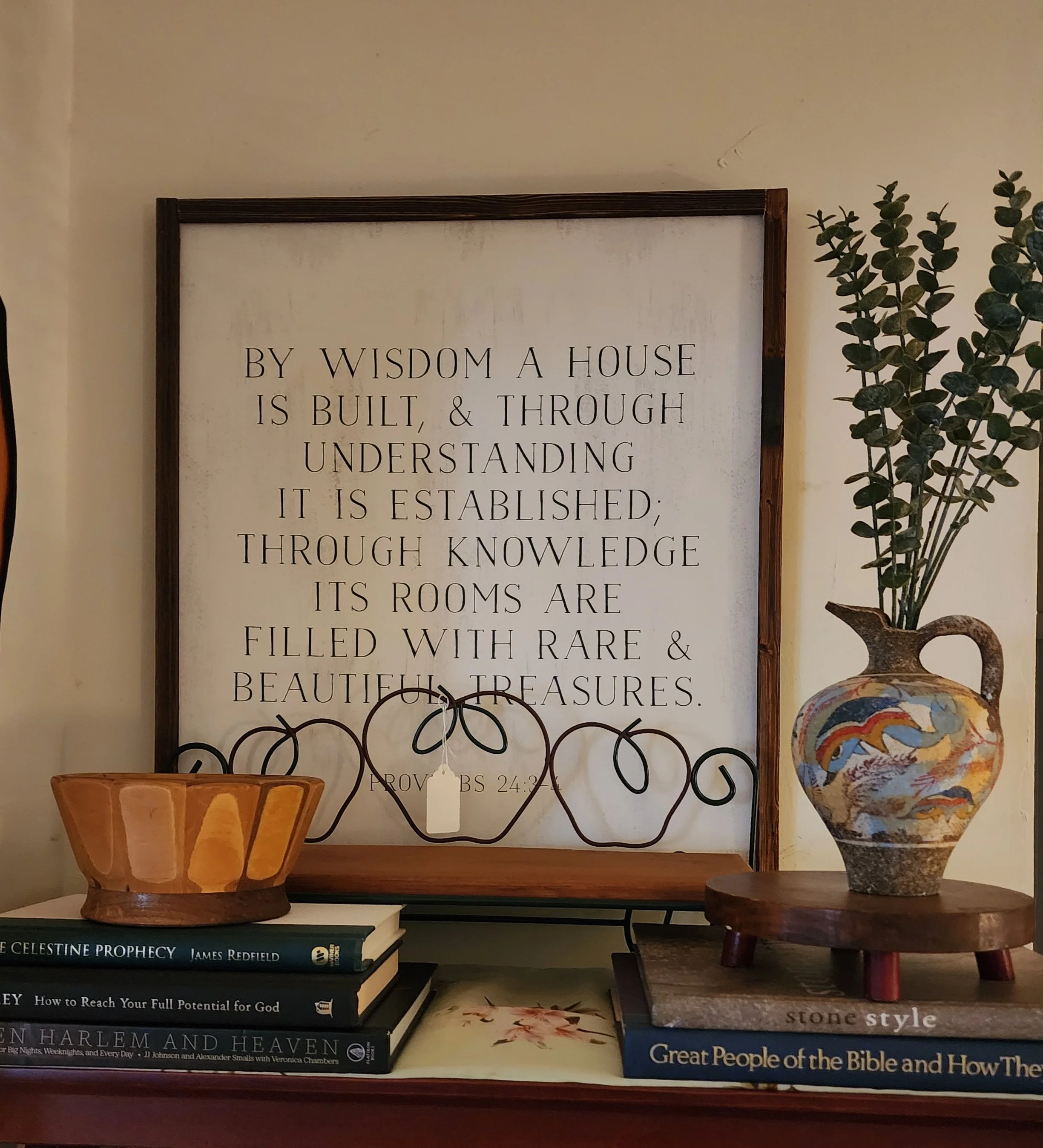 Decorative shelf with books, a ceramic bowl, a small wooden table, a leafy plant in a colorful vase, and a framed quote about wisdom and knowledge.