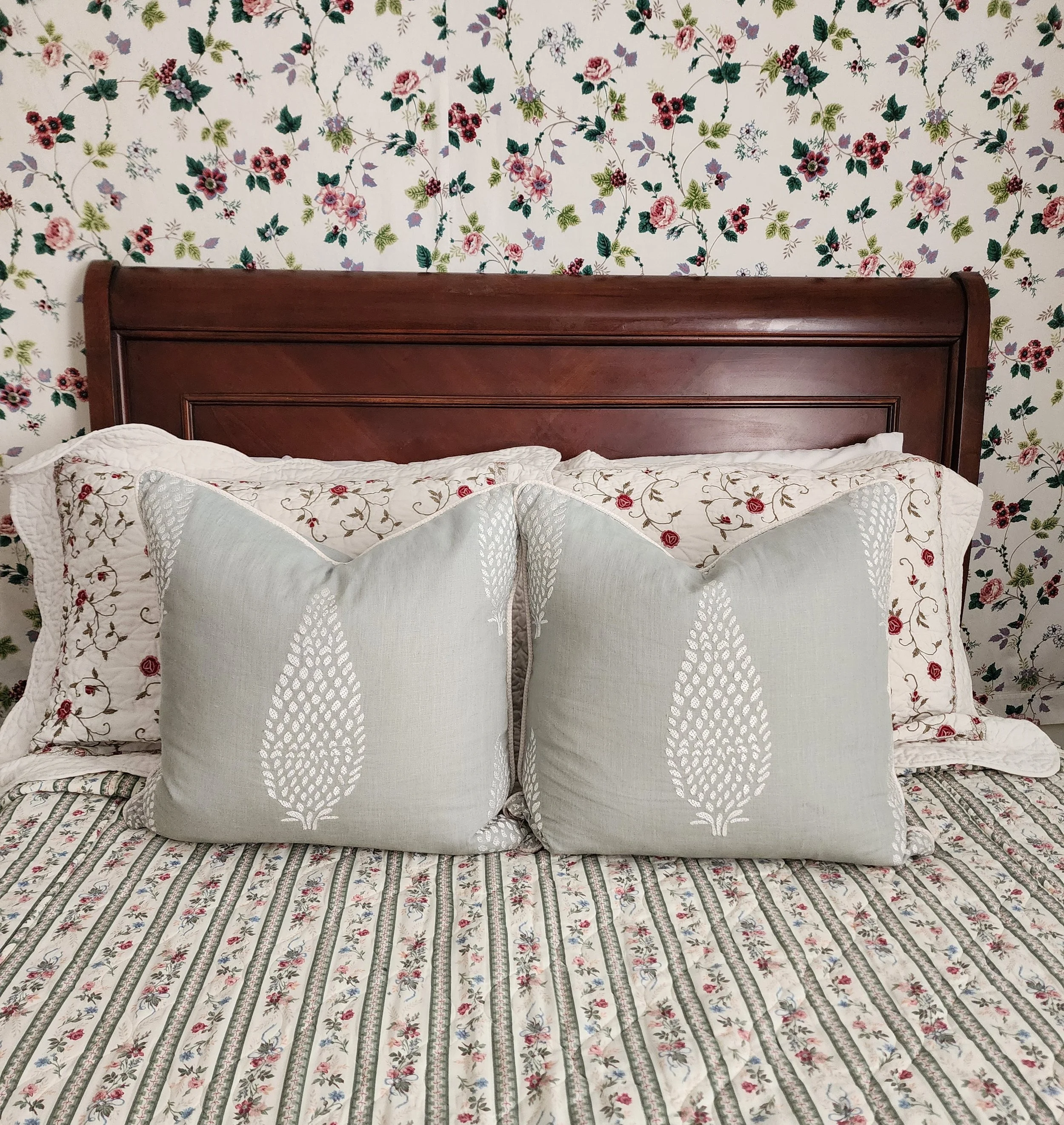 A bed with floral bedding, decorative pillows, and a wooden headboard against a floral wallpaper background.