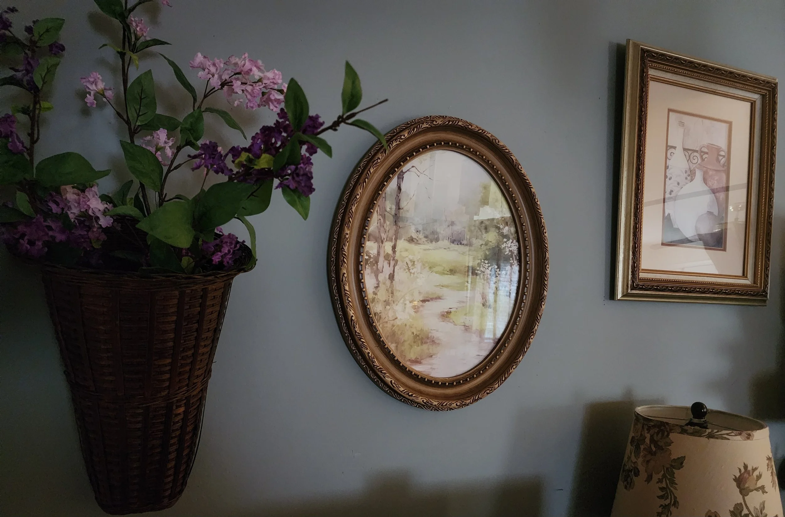 Decorative interior wall with a flower arrangement in a basket, a landscape painting in an ornate oval frame, a framed artwork, and a floral-patterned lampshade.