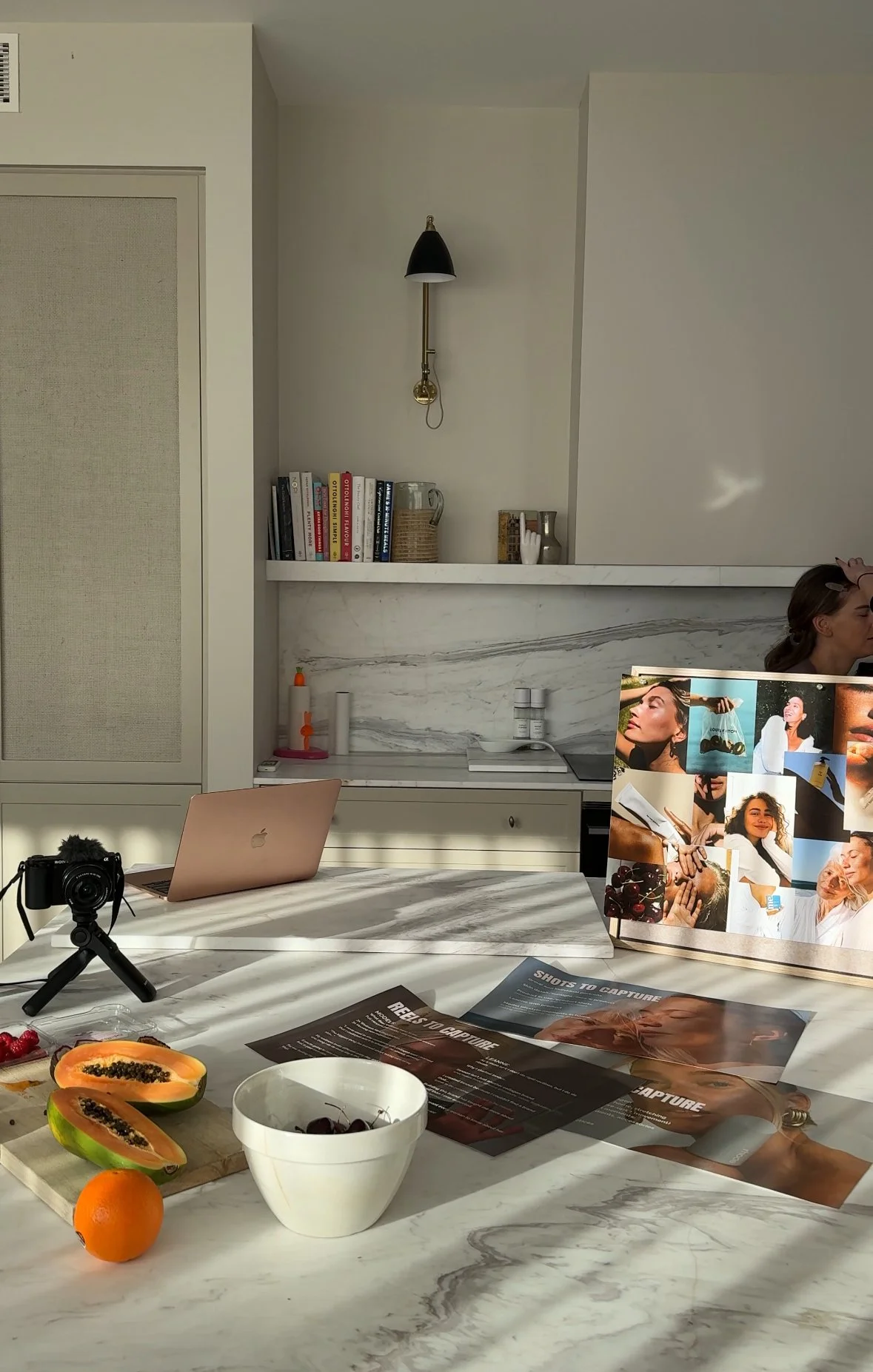 A kitchen with a marble countertop, a white cabinet, a laptop, a camera on a tripod, plates with papaya and orange, a white bowl with cherries, various papers, and a collage of photos on display. Sunlight casts shadows on the counter.