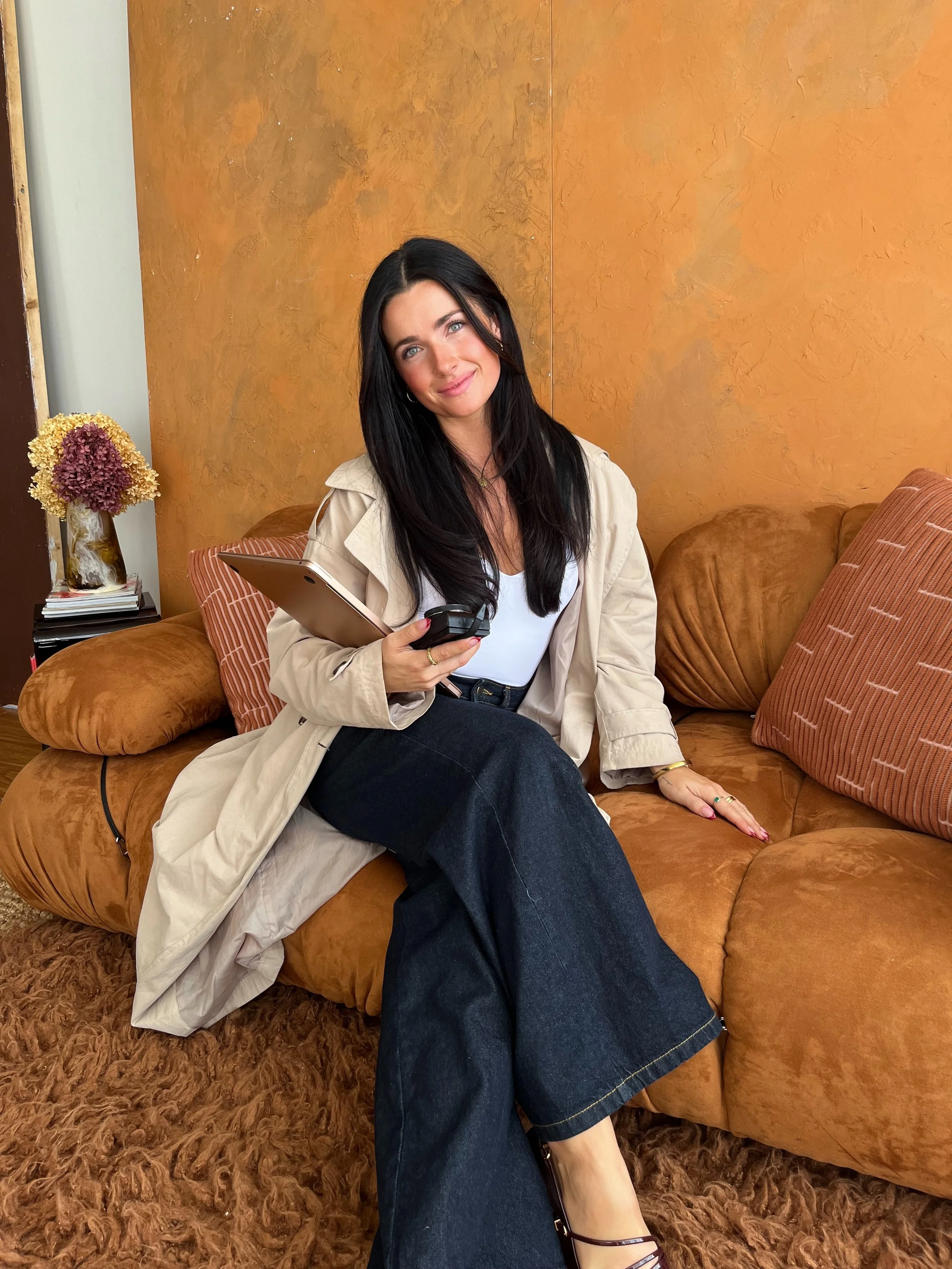 A woman with long black hair, wearing a beige trench coat, white top, and dark wide-leg jeans, is sitting on a brown couch. She is holding a tablet in her right hand and looks at the camera with a slight smile.