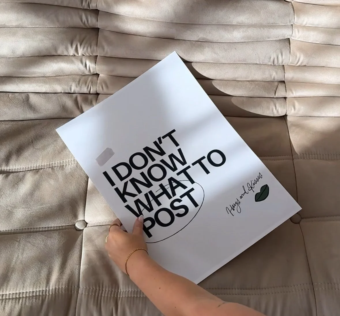 Person holding a white poster with bold black text that reads, 'I DON'T KNOW WHAT TO POST,' on a beige quilted fabric surface.