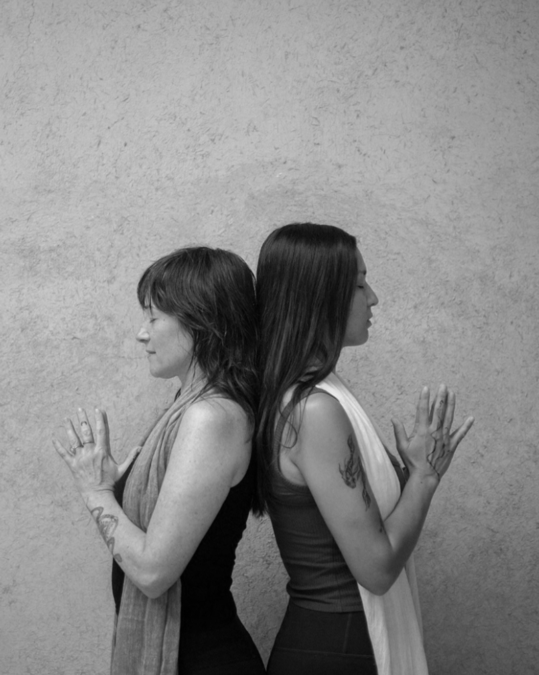 Two women standing back-to-back with closed eyes and clasped hands, against a plain textured wall.