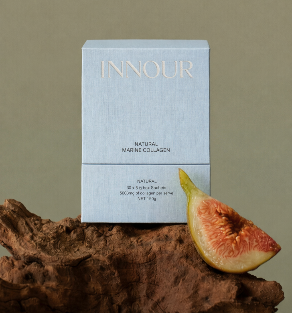 A light blue box of INNOUR Natural Marine Collagen sachets resting on a piece of driftwood with a fresh fig slice in front of it.