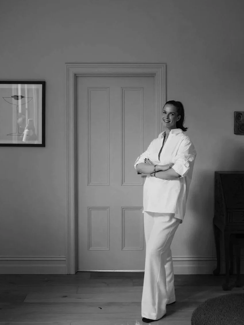 A woman in white clothes standing in a room, smiling and crossing her arms.