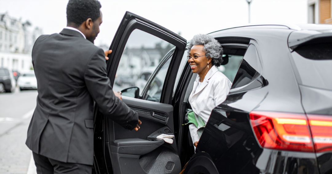 An elderly woman with gray curly hair and glasses smiling as she steps out of a black SUV, assisted by a man in a suit holding the car door open, on a city street with parked cars and buildings in the background.