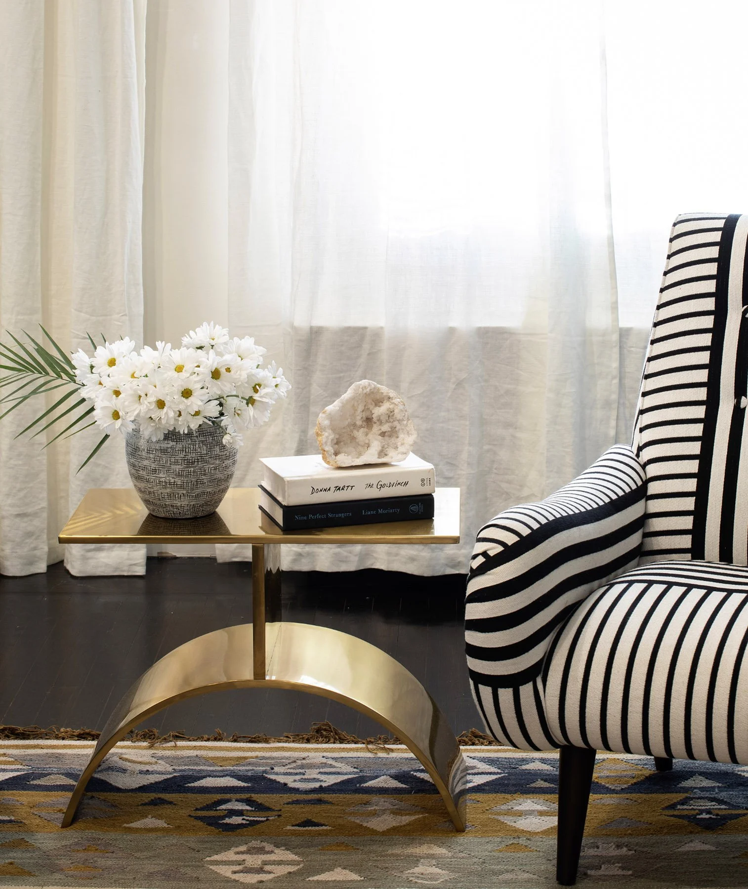 A living room corner with a small gold table holding a vase of white daisies and books with a geode on top. Part of a striped armchair is visible next to the table, and sheer white curtains cover the window in the background.