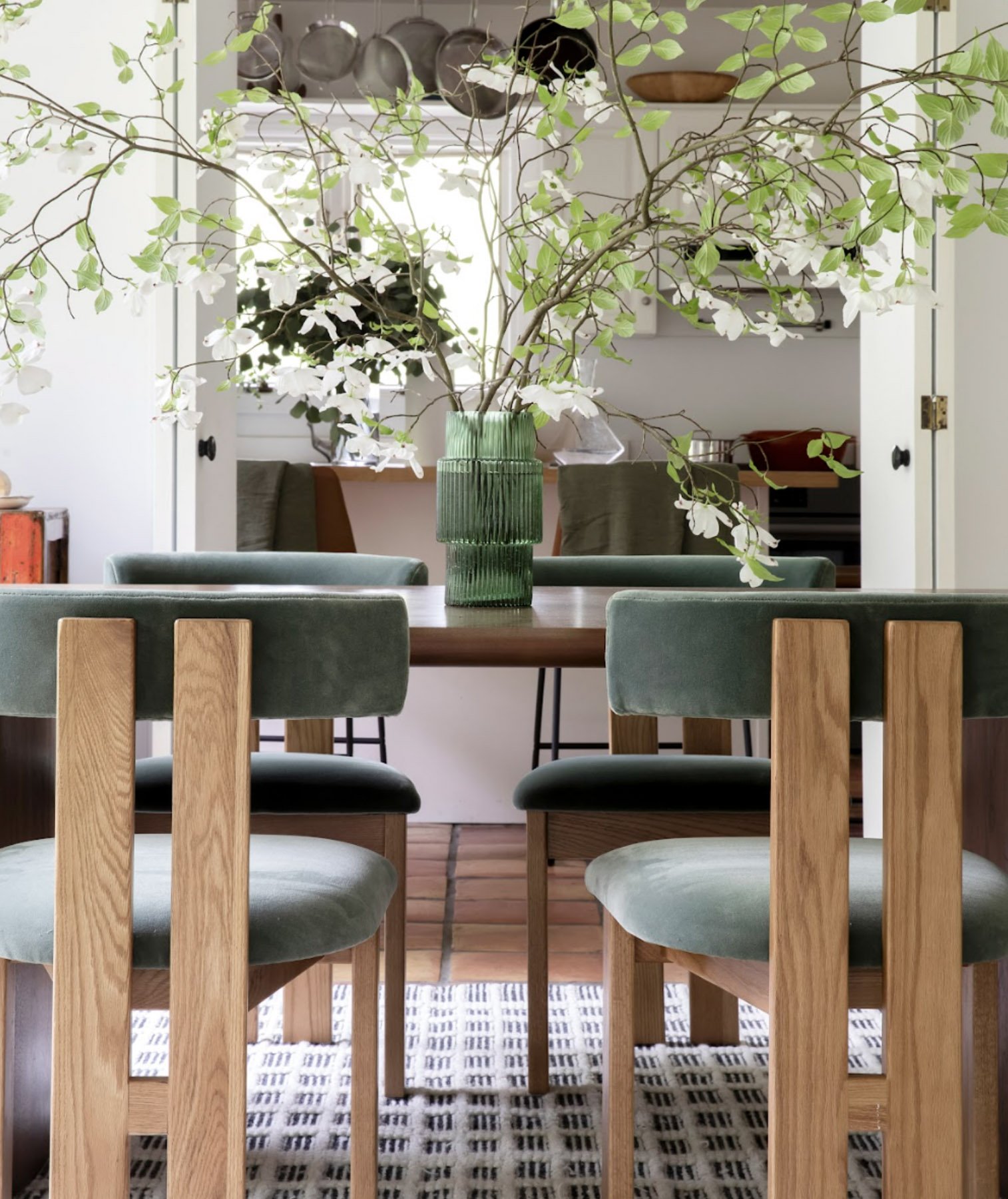 A dining room with a wooden table, green upholstered chairs, and a large green vase with white flowering branches as a centerpiece. There is a patterned rug underneath the table, and shelves with kitchenware in the background.