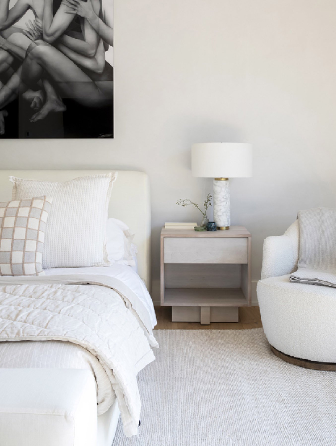 A bedroom with a white bed, pillows, a beige nightstand with a marble table lamp, a small plant, and a stack of books. There is a white armchair with a blanket, and a black-and-white artwork on the wall.