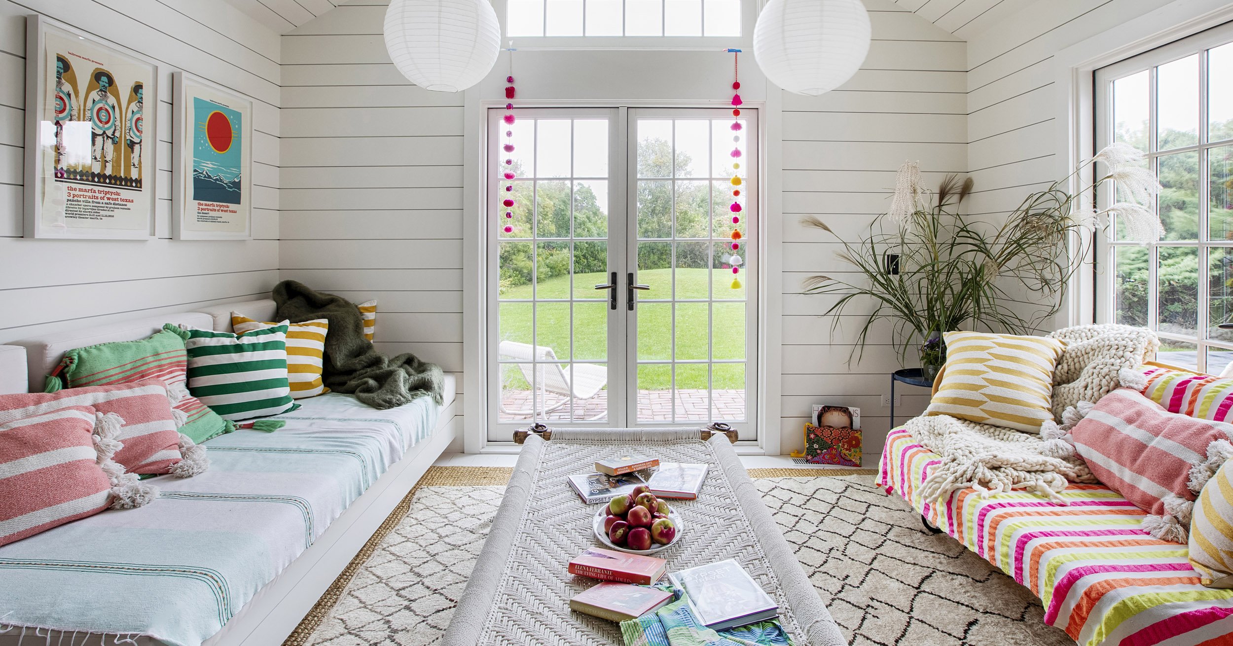 Bright living room with white shiplap walls, two large sofas with colorful striped and solid pillows, a woven coffee table with books and apples, gallery wall art, large windows and glass double doors opening to a green outdoor space, and hanging pap
