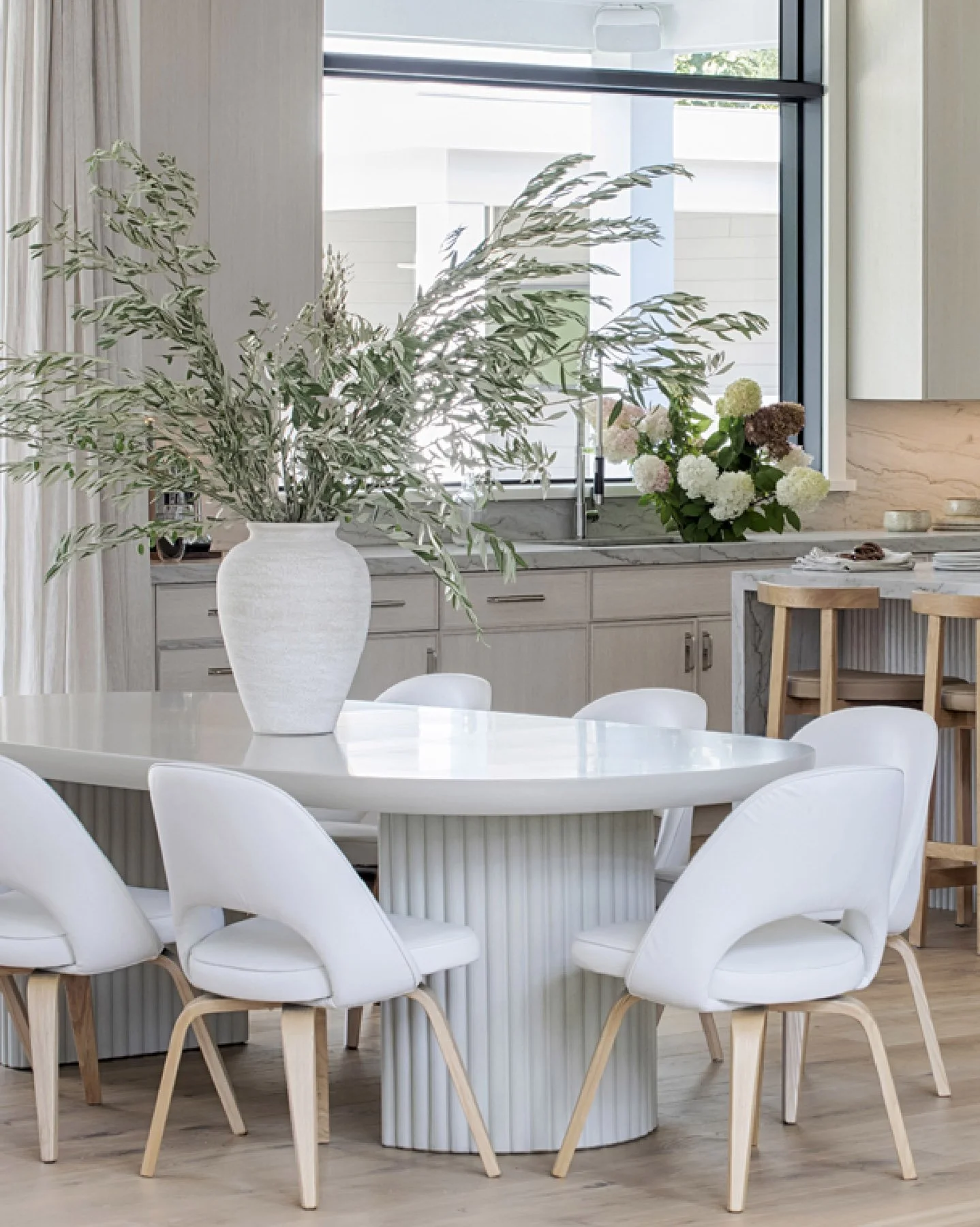 Modern Hamptons dining area with a round white table, white chairs, a large vase with green and white foliage, and a window with flowers in a vase on the window sill.