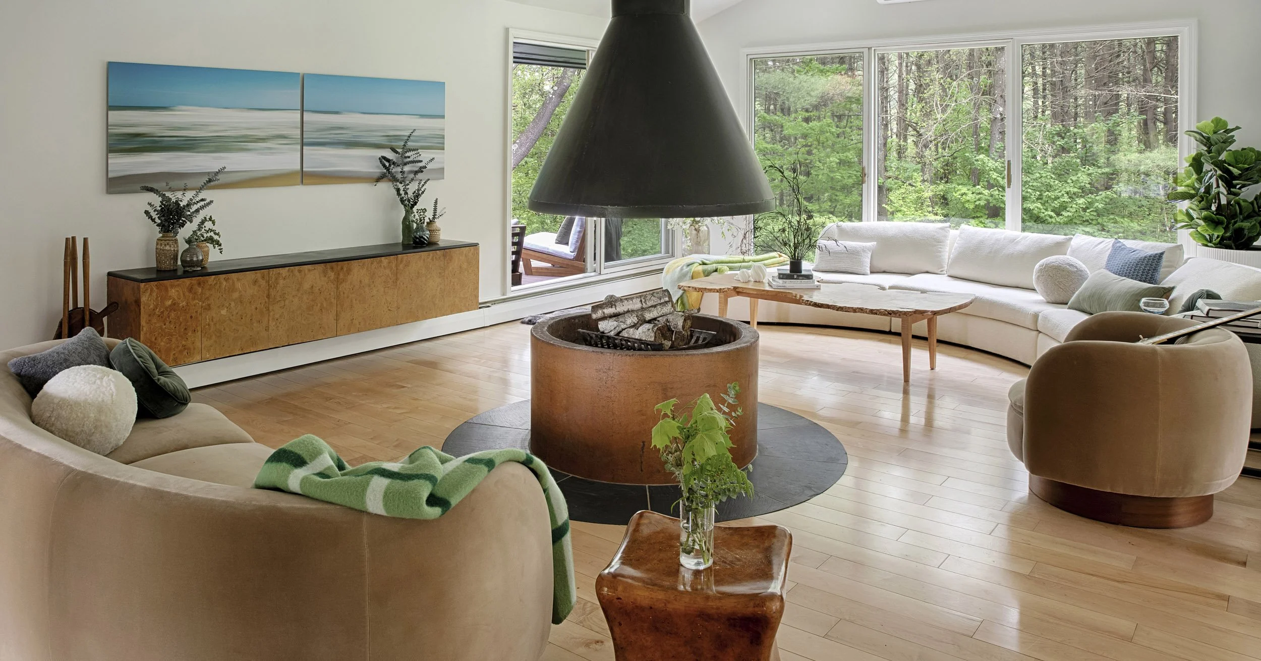 Living room with wood floor, beige and white sofas, large windows overlooking trees, and a central fireplace with logs, under a large black hood. Decor includes plants, pillows, artwork, and a wooden side table.