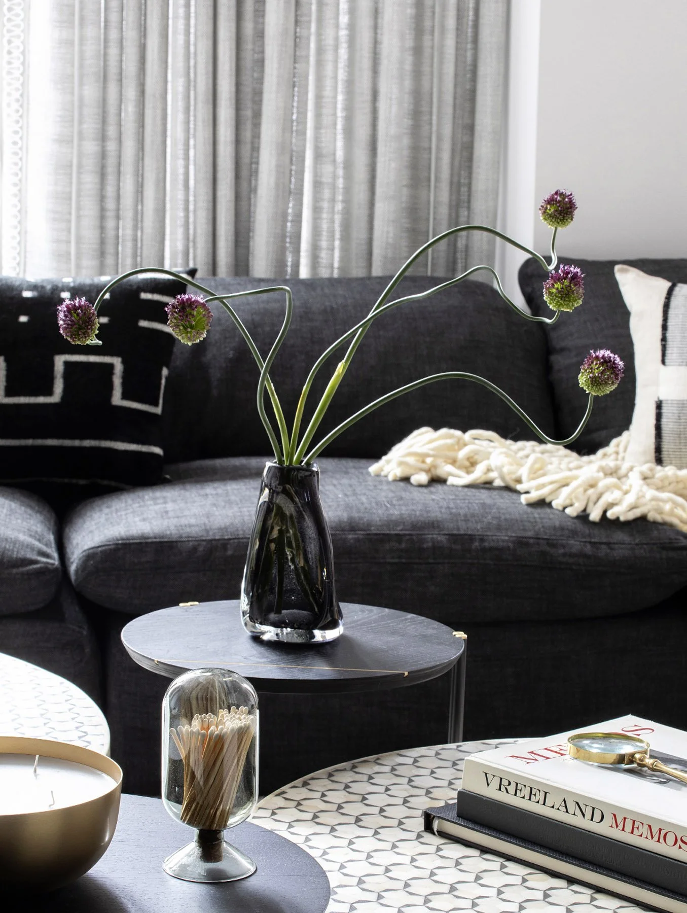 A living room with a dark gray sofa, pillows, a throw blanket, and curtains, featuring a decorative black vase with curly chive flowers on a small round table.