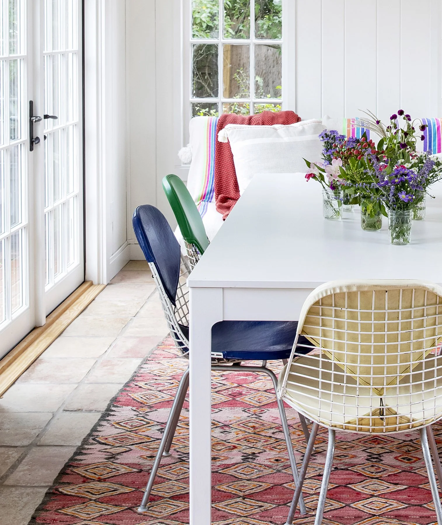 A dining area with a white table, colorful wire chairs, and a floral centerpiece, adjacent to a white sofa with pillows and blankets, near glass doors and windows revealing a garden outside.