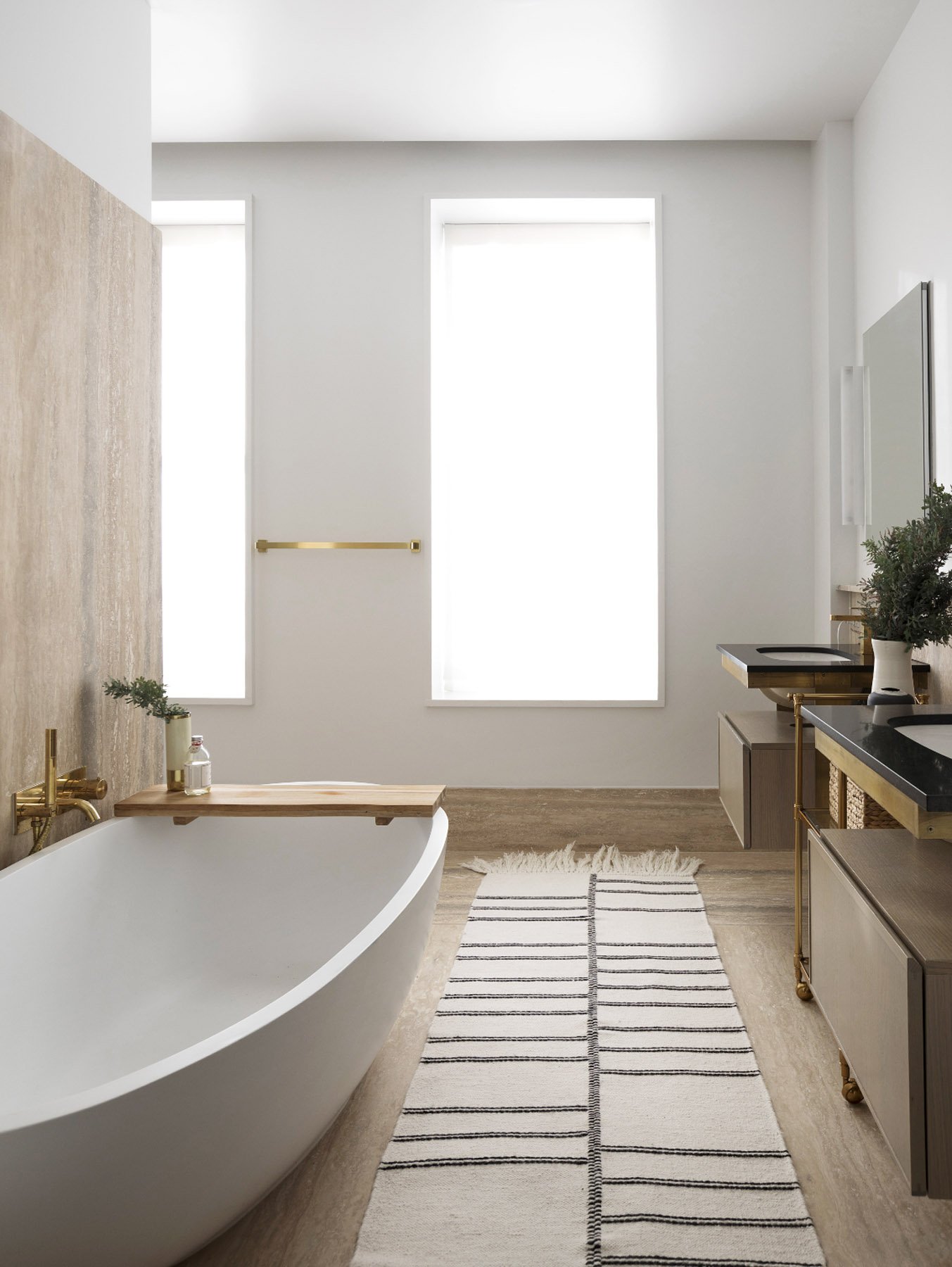 Modern bathroom with a white bathtub, wooden countertop, and double sinks with black countertops. Two tall frosted windows let in light, and a striped rug lies on the wooden floor.