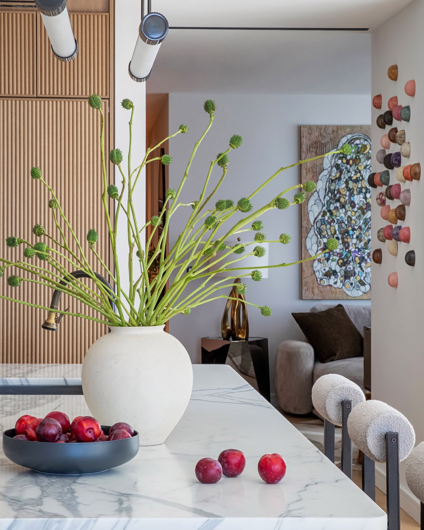 A modern living room with a white marble dining table holding a white vase with green flower stems and a bowl of red apples. In the background, there are abstract art and a beige couch with a brown pillow, with colorful small cups lined on the wall.