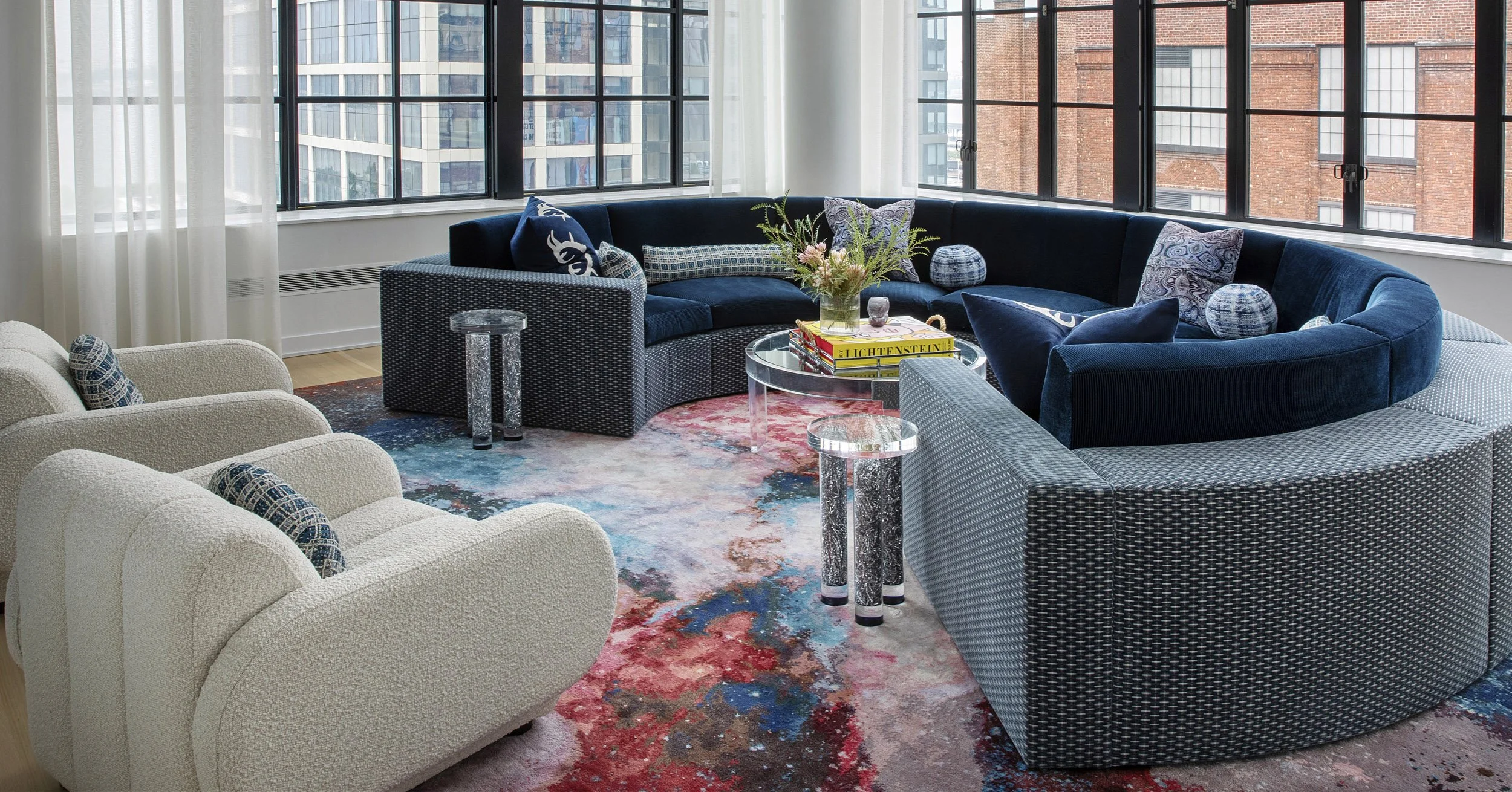 Modern living room with large windows, curved navy blue velvet sectional sofa, white textured armchairs, glass coffee table with books and a floral arrangement, abstract multicolored area rug, cityscape view outside windows.