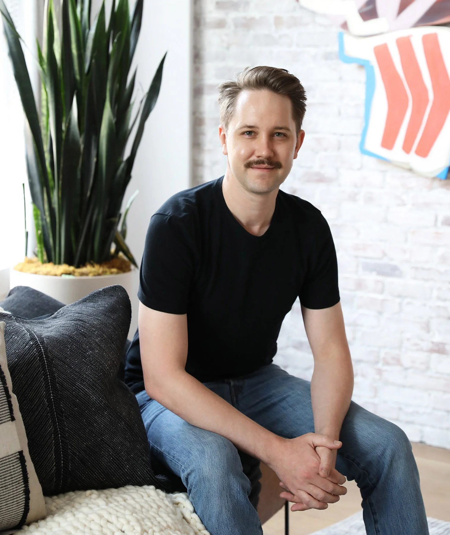 Tyson Ness, founder of Studio Ness, sitting on a couch in a modern living room with a brick wall background and abstract artwork.