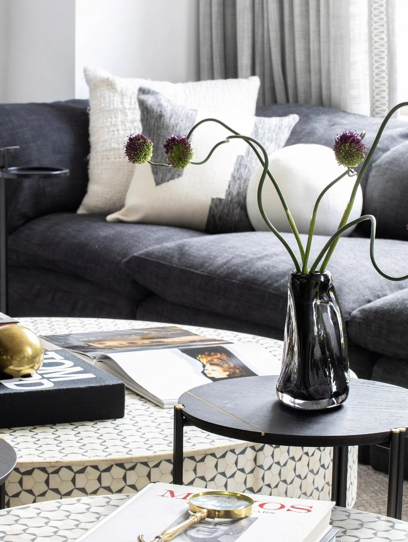 Living room with a gray sofa, white and gray pillows, a black vase with purple flowers on a small round black table, an open magazine, and contemporary decor.