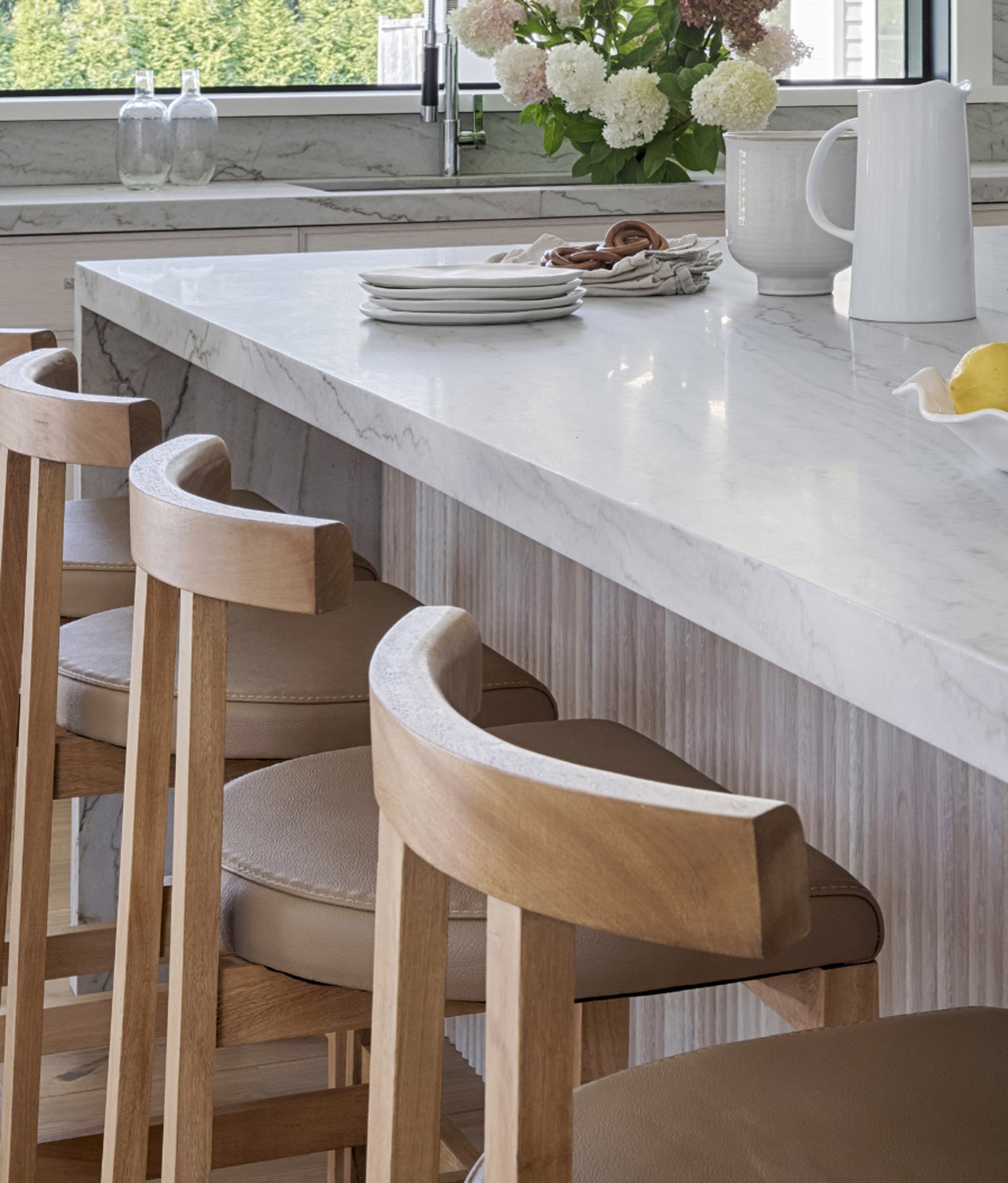 A modern kitchen with a white marble countertop, light wooden chairs, and a vase of white hydrangea flowers by the window.