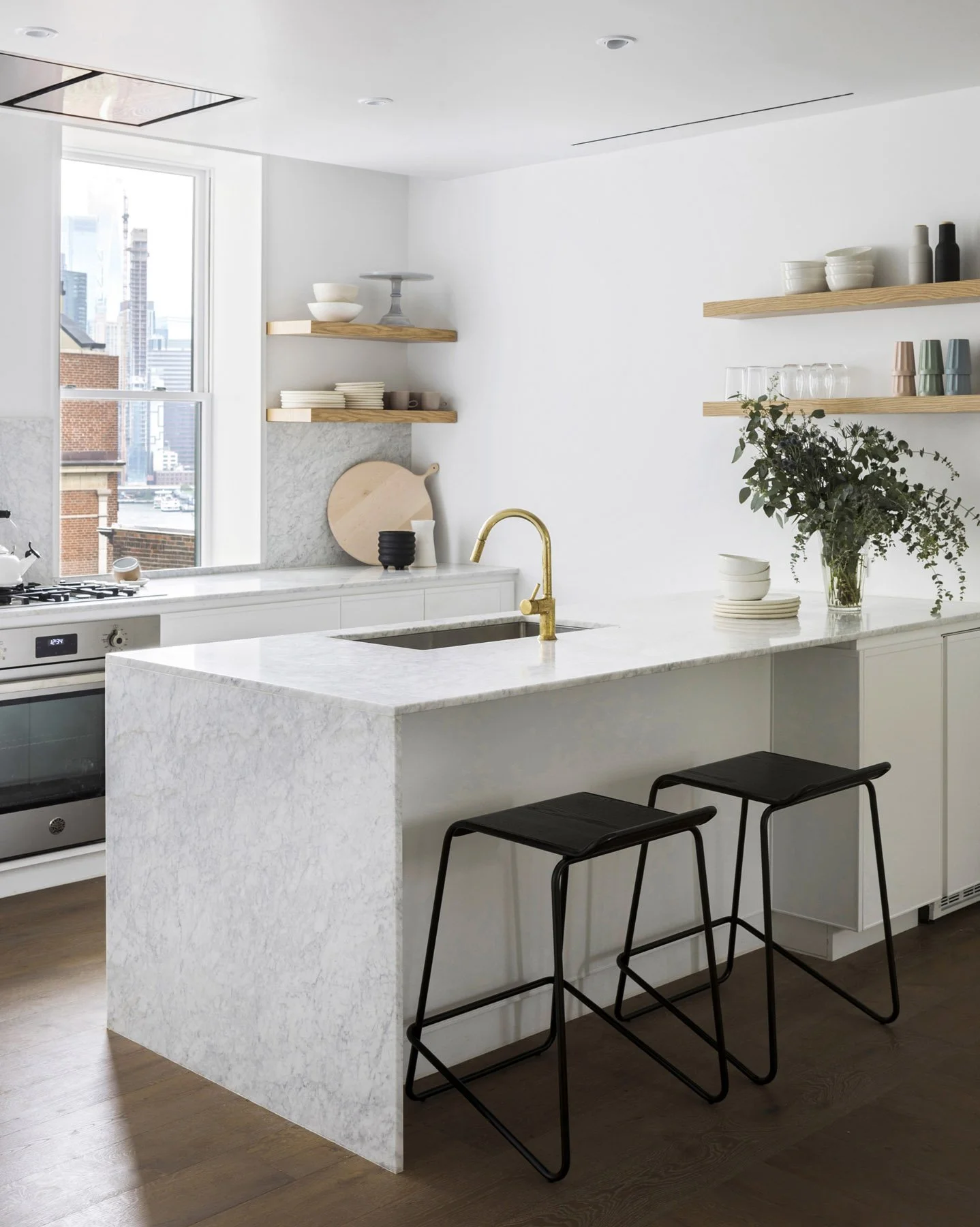 Modern kitchen with white marble island, black barstools, open wooden shelves with dishes and plants, and city skyline view through large window.