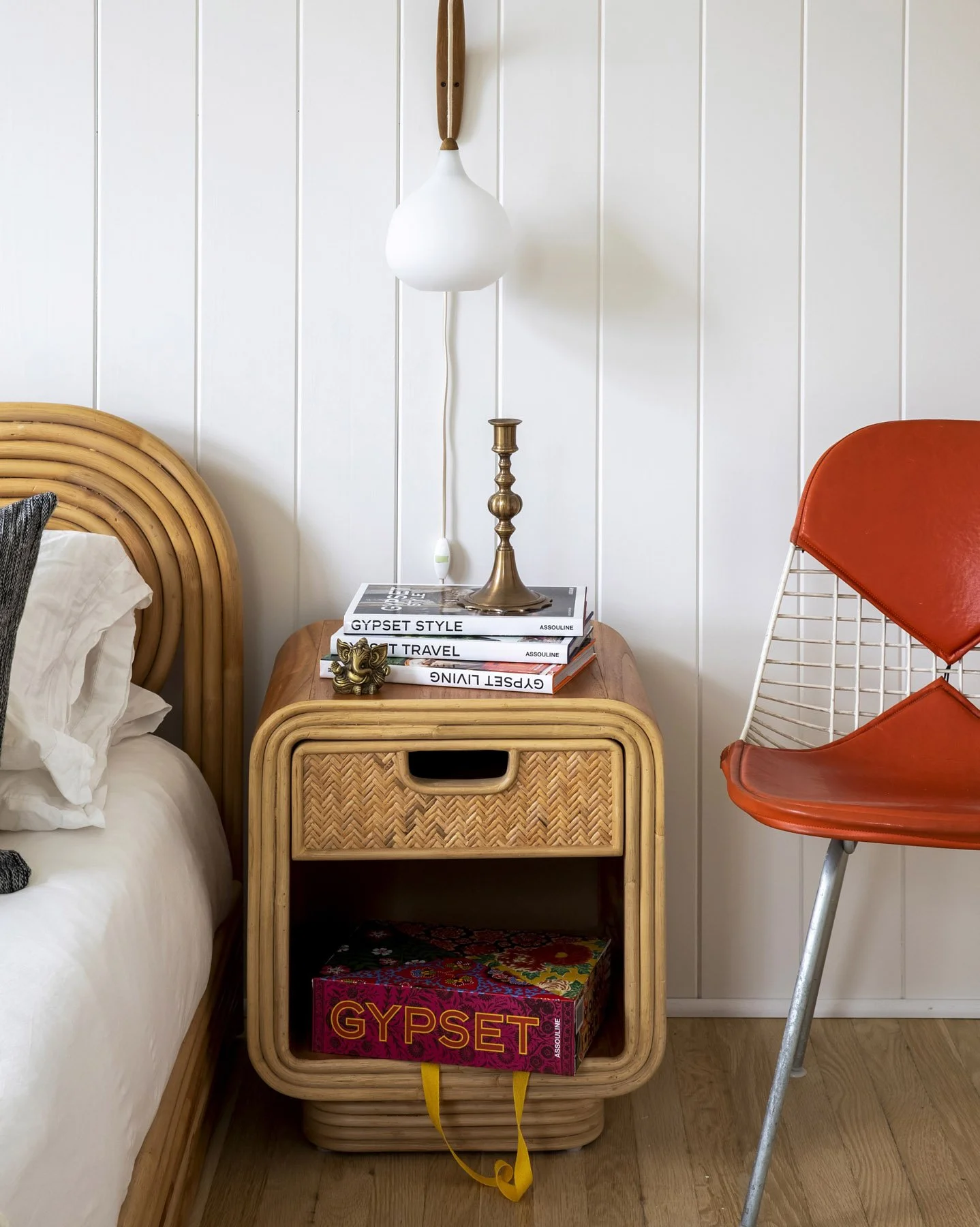 A nightstand with a rattan design, topped with a stack of books including 'Gypsy Style' and 'Great Living'; a brass candlestick, a small decorative lion figure, and a white pendant lamp hanging above. To its right is a retro orange chair with a wireframe backrest, and a portion of a bed with a rattan headboard is visible to the left.
