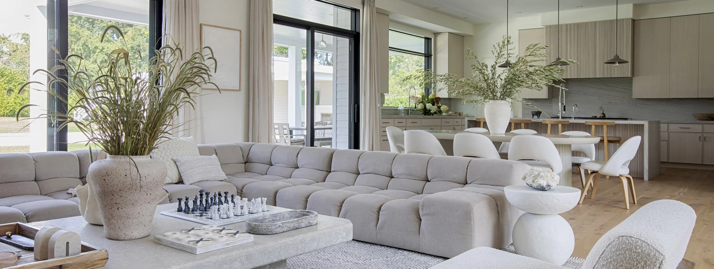 Living room with large sectional sofa, potted plants, and a kitchen area with a dining table and white chairs.