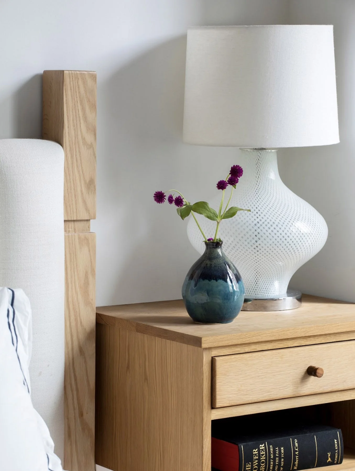 A wooden bedside table with a black and blue vase holding purple flowers, a white table lamp, and a few books underneath.
