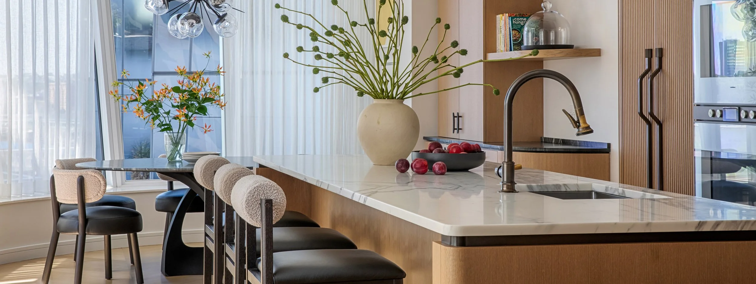 Modern kitchen with a marble island, large vase with green plants, bowl of red and purple plums, dining table with a clear glass vase containing orange flowers, and soft natural light from large windows with sheer curtains.