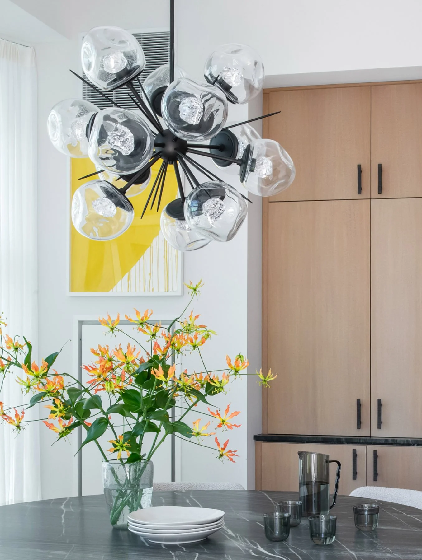 Interior of a dining room with a bowl of plates, a vase of orange and yellow flowers, and a glass pitcher with glasses on a dark stone table. A modern chandelier with glass globes hangs from the ceiling, and there is a yellow abstract painting on the wall.