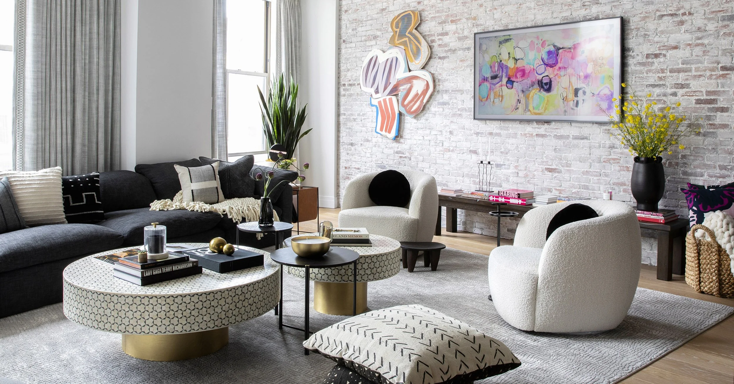 Modern living room with white brick wall, black and white furniture, colorful wall art, yellow flowers in a black vase, and decorative items on coffee tables.