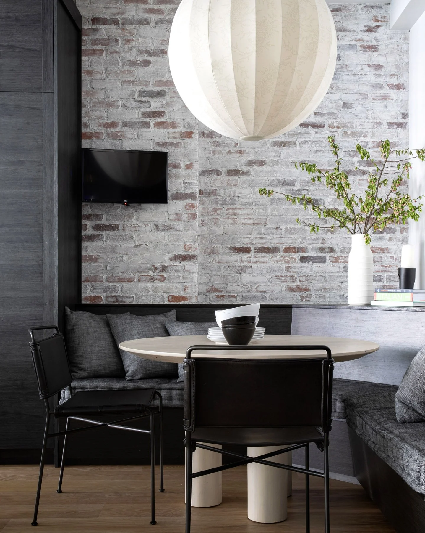 Interior of a modern dining nook with a round table, black and gray chairs, a gray stone bench with pillows, a white vase with branches, a wall-mounted TV, and a large paper lantern ceiling light.