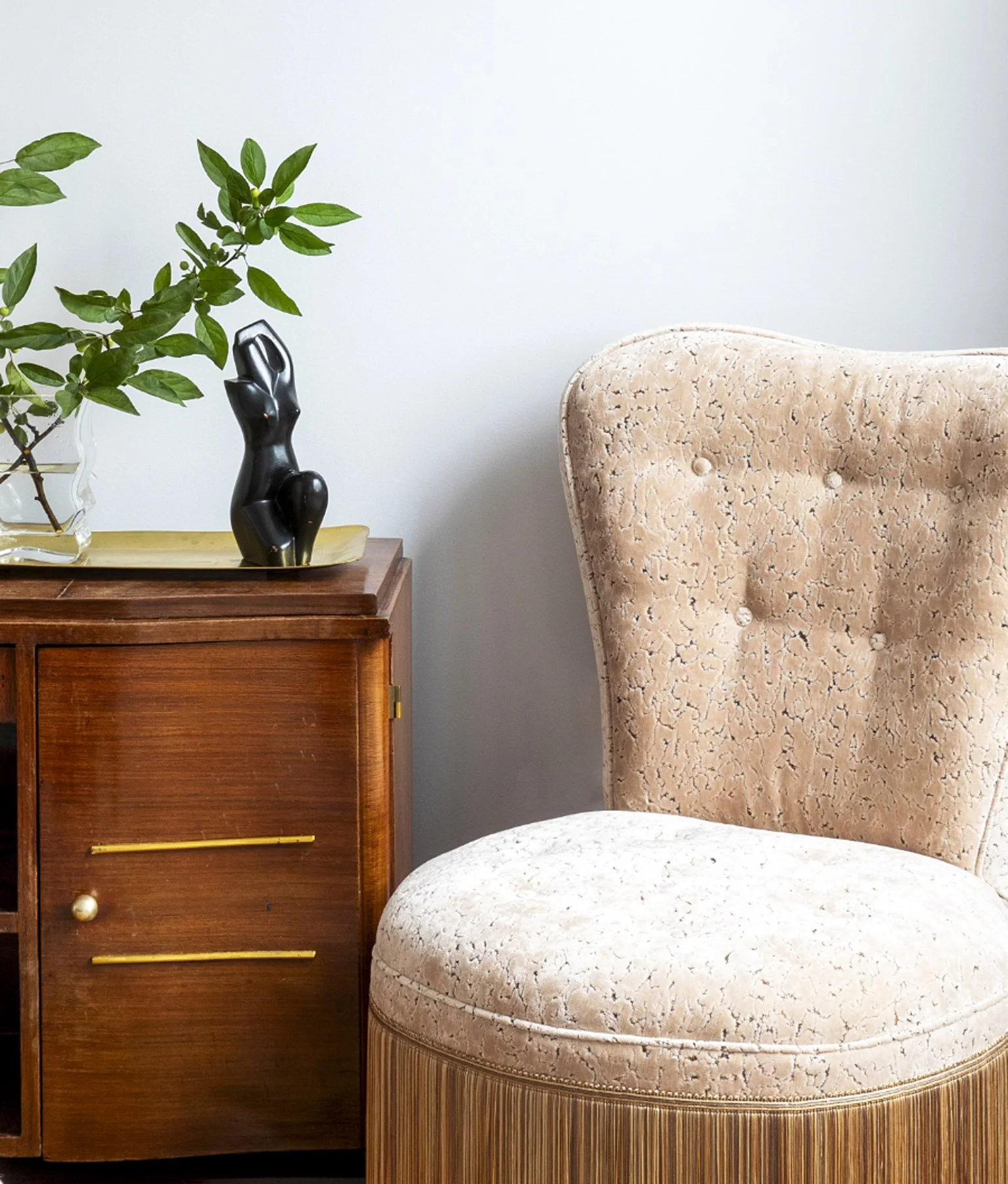 A beige vintage armchair with textured upholstery next to a wooden cabinet, decorated with a black abstract sculpture and a potted plant with green leaves.