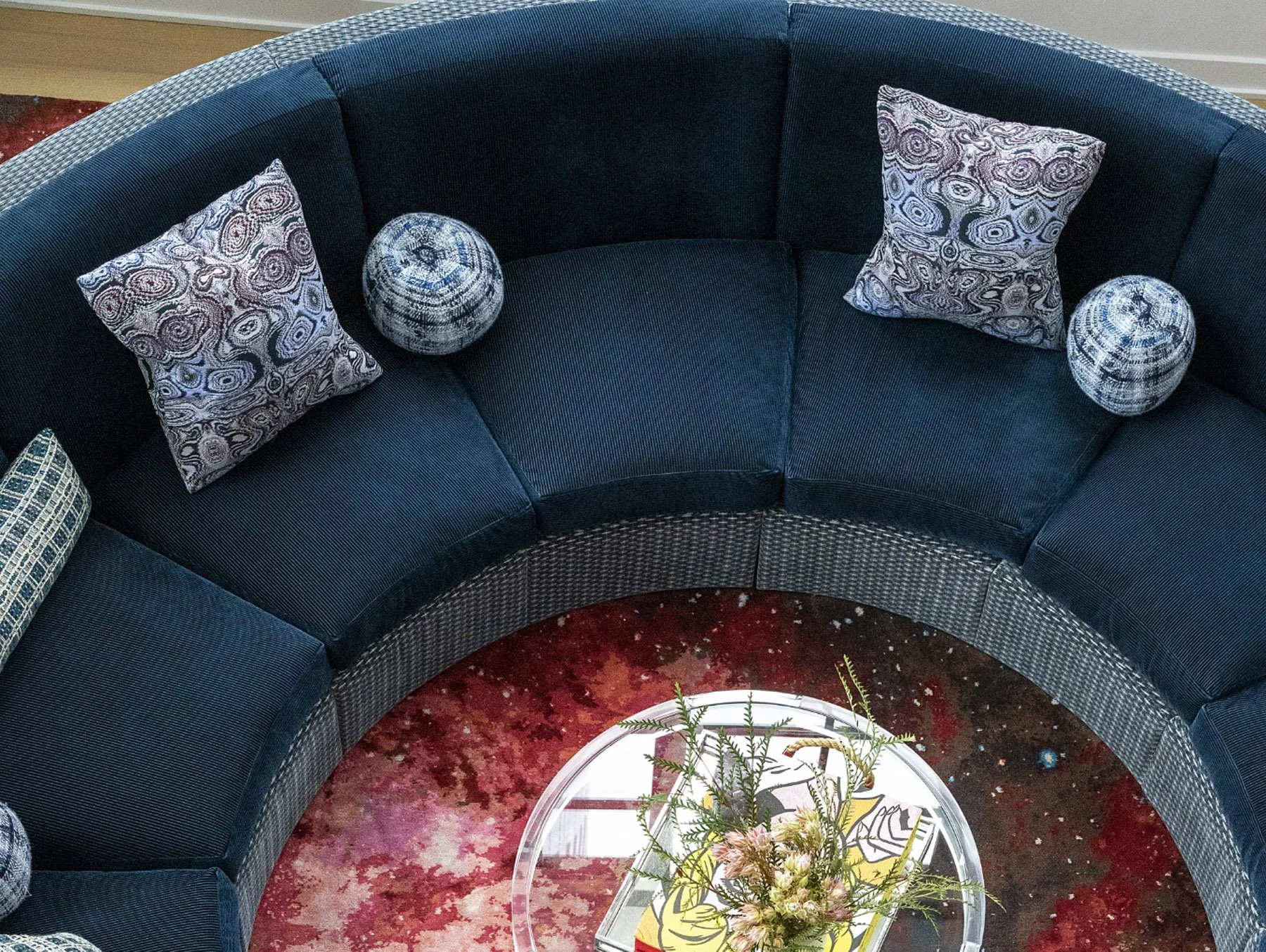 A curved navy blue sofa with patterned pillows and spherical decorative objects, a glass-topped coffee table with a floral arrangement, set on a red abstract rug.
